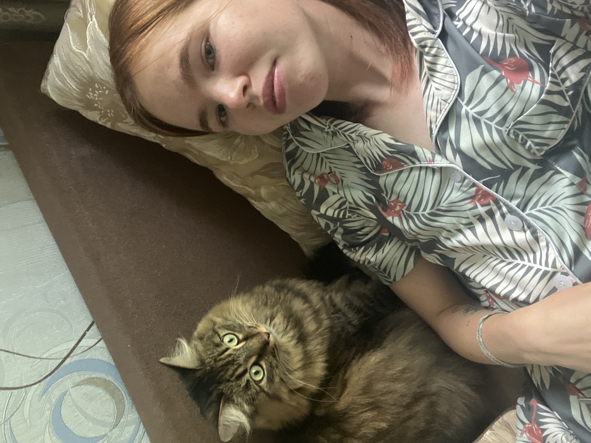 A young woman with reddish-blonde hair, wearing pajamas with a tropical leaf and flamingo print, is lying on a sofa with her cat. The cat, a fluffy tabby with green eyes, is sitting next to her. The sofa is covered in a brown fabric, and a patterned pillow is visible behind the woman. The background shows a wall with light blue wallpaper featuring a circular design and a thin brown line. The scene appears to be indoors, likely in a living room. No specific details suggest a location in Chaikovsky, Russia, beyond the provided context.