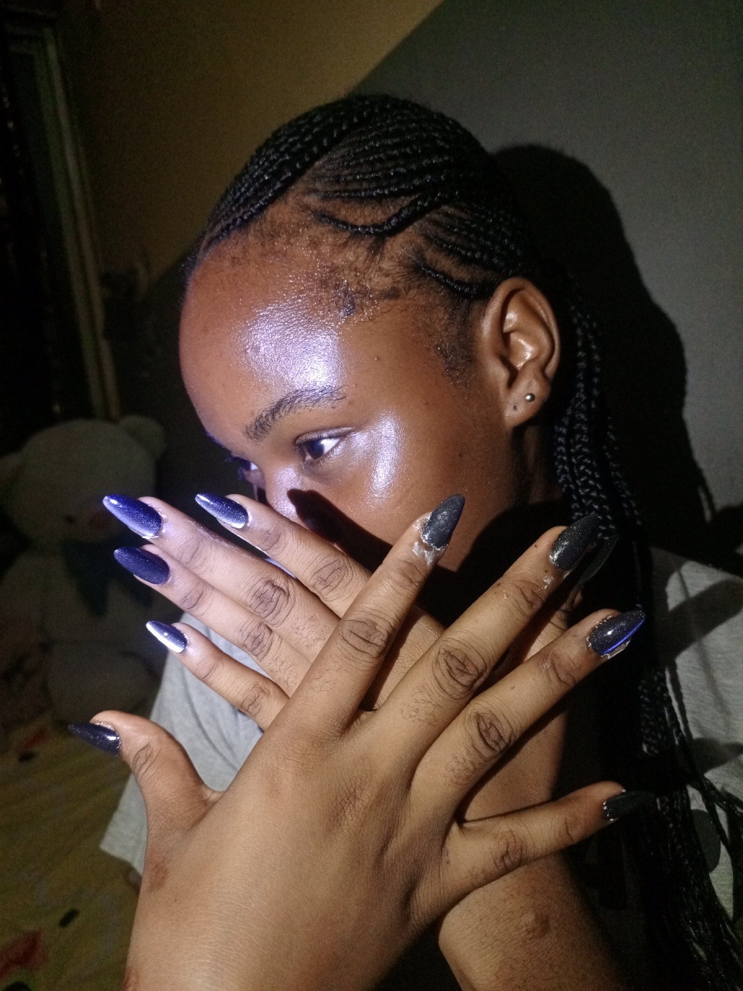A young, dark-skinned individual is depicted indoors. The person has cornrow braids and pierced ears. The focus is on their hands, which are crossed in front of their face and display long, pointed nails with a dark glitter finish and a lighter tip on some fingers. There is a teddy bear in the background. The location of the image is unknown and is not identifiable in the city of Keffi, Nigeria.
