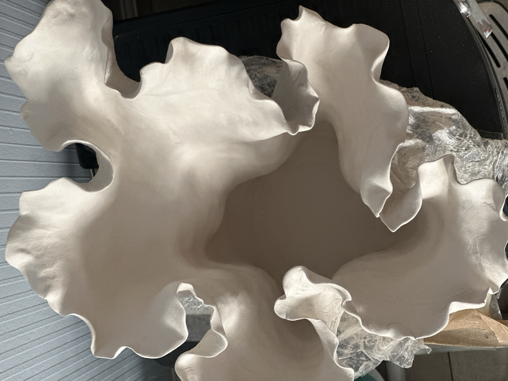 The image shows a close-up, overhead view of a white, irregularly shaped ceramic bowl with ruffled edges. The bowl is placed on a dark surface, possibly a table or counter. Some crumpled, translucent plastic material is visible beneath and around the edges of the bowl. In the background, a striped wall or surface is partially visible. There are no people, actions, or interactions depicted in the image. The location in the city of Nevinnomyssk, Russia, is not identifiable from the visual information provided in the image.