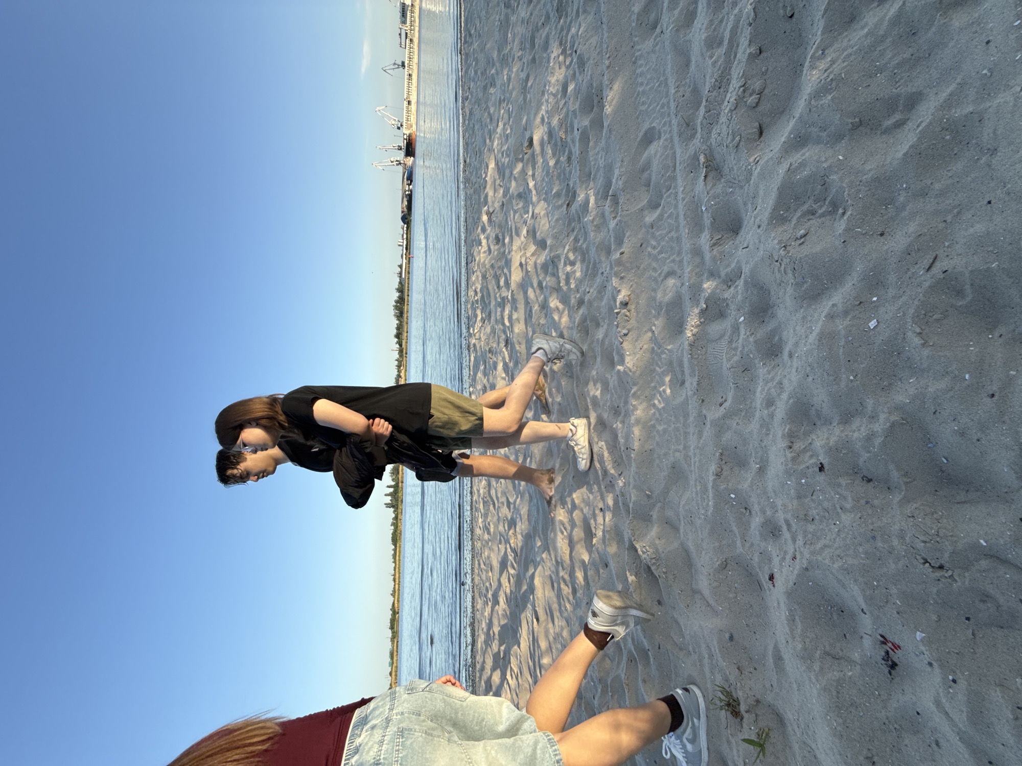 The photograph depicts a beach scene, possibly in the vicinity of Rostov-on-Don, Russia, given the presence of harbor cranes in the background. Two figures are partially visible in the foreground; one is wearing a maroon shirt with denim shorts, and the other is wearing a black shirt with green shorts. The figures are standing on a sandy beach, and water is visible in the background between the beach and the skyline featuring cranes.