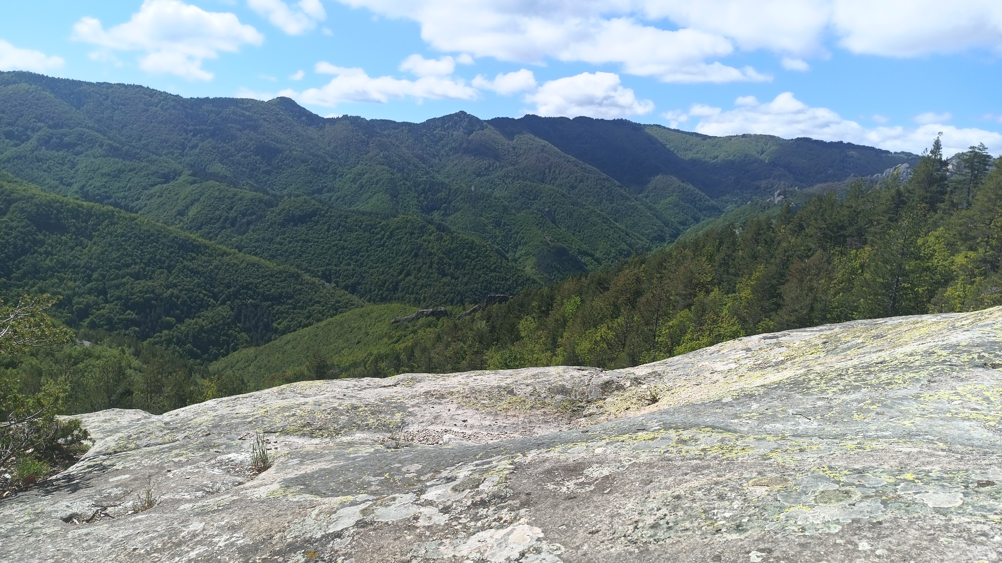 The image shows a landscape view from a rocky outcrop in Sini Vrah, Bulgaria. The foreground consists of a textured, light-colored rock surface with patches of moss and lichen. In the midground, there's a dense forest covering rolling hills and valleys, mostly green with varying shades. In the background, there is a ridgeline of mountains covered in forest. The sky is blue with scattered white clouds. No people are visible in the image.