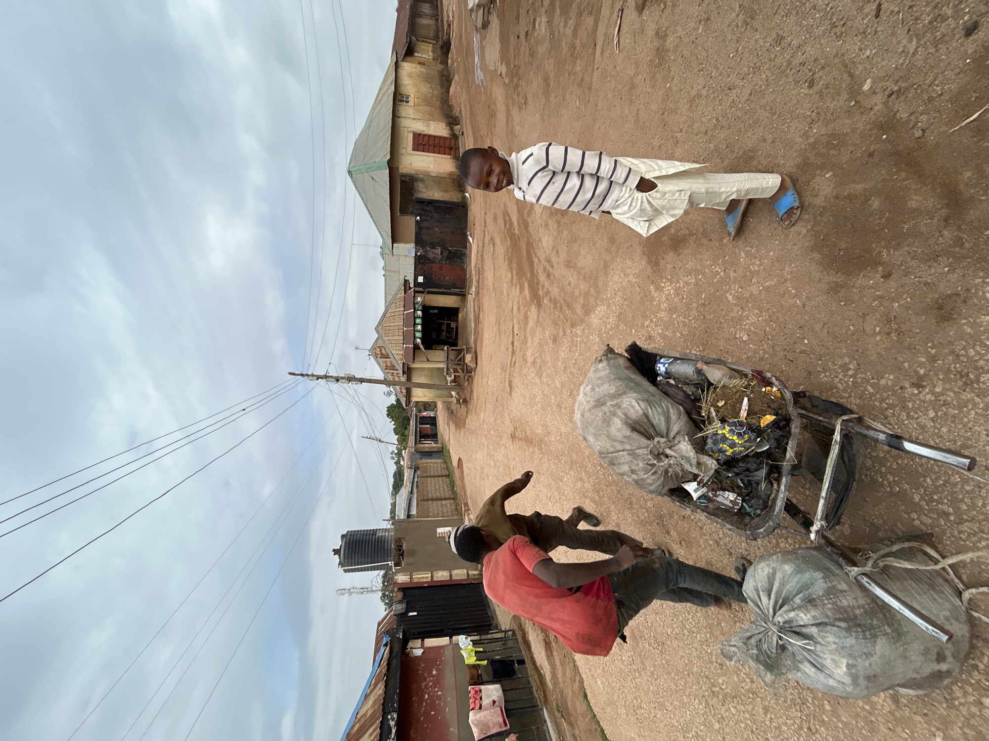 The image, oriented with the sky towards the left, depicts a scene on a dirt road likely within a less affluent area of Jos, Nigeria. A boy wearing a striped white and black sweater and light-colored pants is standing and facing the camera. He is wearing blue sandals. In the foreground, a man in a red shirt is struggling with a wheelbarrow filled with trash and two large, gray bags of refuse. The wheelbarrow is filled with visible plastic containers and discarded items. A second man appears to be playfully wrestling with the man pushing the wheelbarrow. The surrounding buildings are simple, single-story structures with corrugated metal roofs. Overhead are numerous power lines. The sky is overcast.