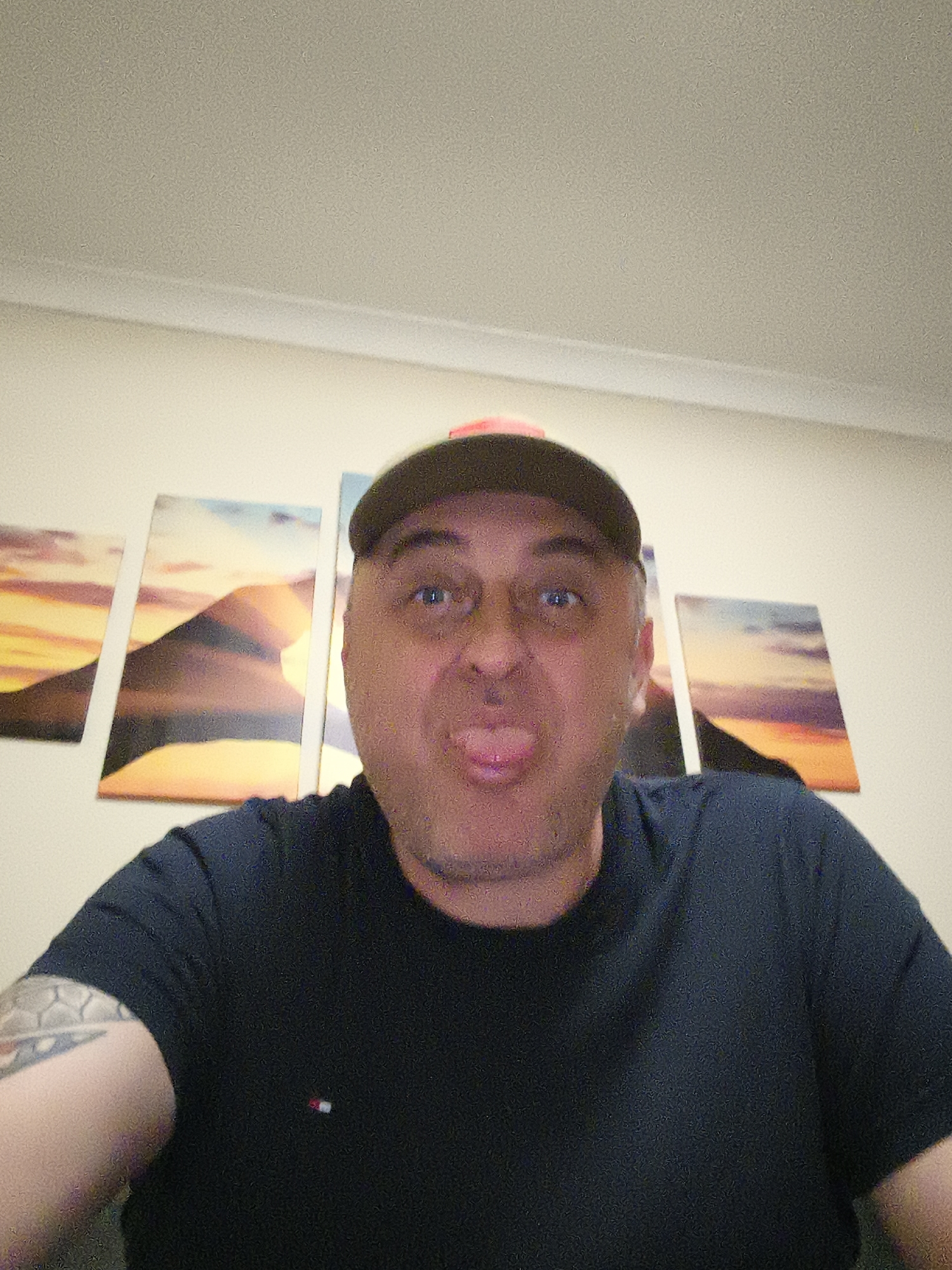 A man, appearing to be in his middle age, is the central subject of this close-up, indoor photograph. He is looking directly at the camera with a playful expression, sticking his tongue out. He wears a dark, possibly navy or black, short-sleeved t-shirt, which features a small rectangular logo resembling the Tommy Hilfiger flag (red, white, and blue) on the left side of his chest. A tattoo with geometric or patterned designs is visible on his left bicep/shoulder. He is also wearing a dark baseball cap, with an olive green or khaki brim and a subtle hint of a reddish or orange color on its crown.
The setting is an interior space, likely a room within a residence in Huntingdonshire, United Kingdom. Behind the man is a plain, light-colored wall. Above the wall, a white crown molding connects to a textured, light-colored ceiling. Four distinct art pieces, possibly canvas prints, are hung horizontally on the wall behind him. These artworks depict abstract landscapes, featuring what appear to be mountains or dunes against skies rendered in warm tones of orange and yellow transitioning into cool blues and purples, suggesting a sunrise or sunset. The lighting in the room appears to be artificial overhead light. The photograph has a slight grainy texture throughout.