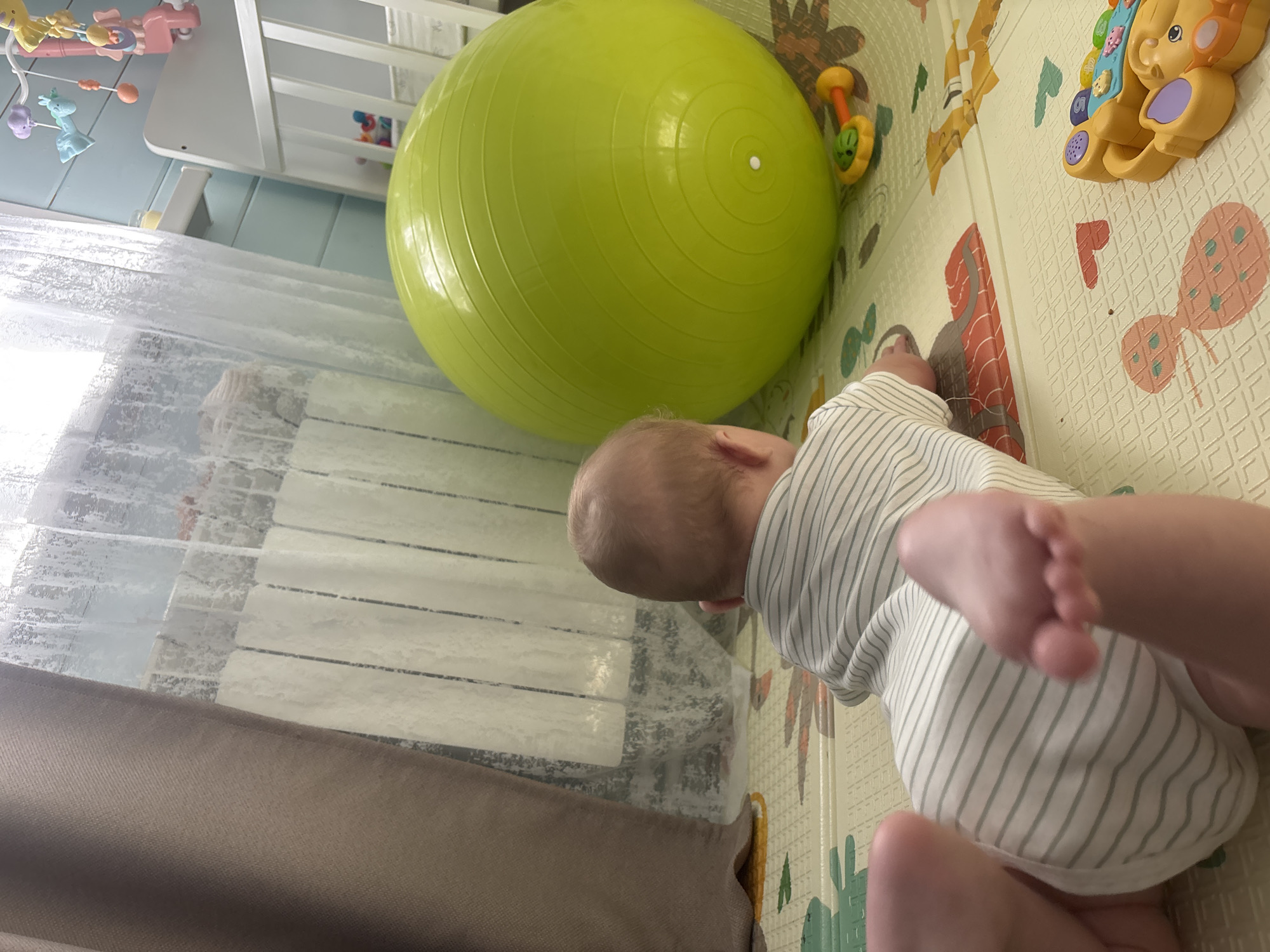 A baby lies on a play mat next to a large green exercise ball and assorted toys in an indoor setting in Раздолье, Russia. The baby is wearing a white and green striped onesie. A crib and a window with sheer curtains are visible in the background.
