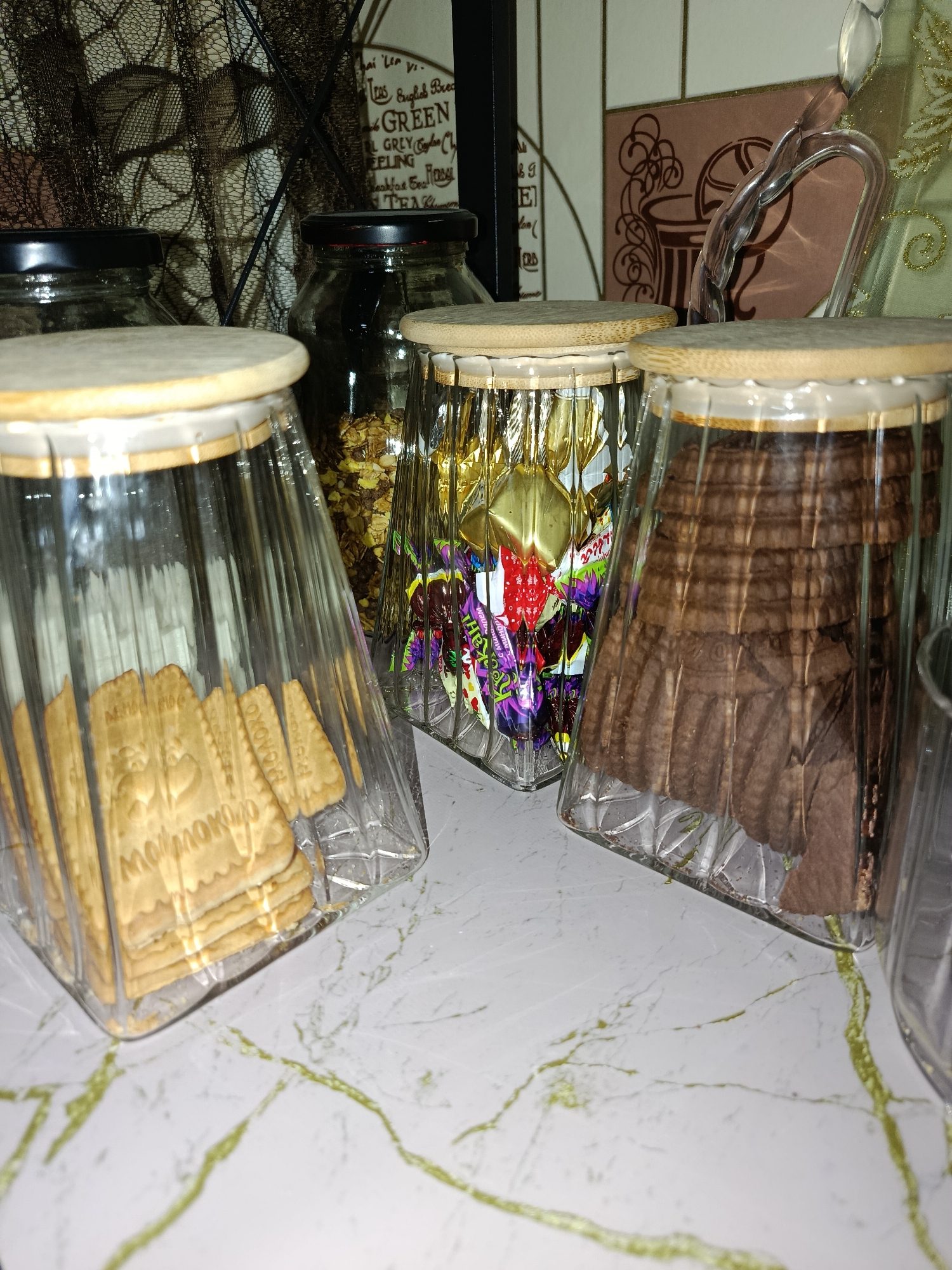 In a close-up, low-angle shot, three glass jars with light wood lids are arranged on a marble countertop. The jar on the left contains stacks of tan-colored biscuits, each imprinted with decorative patterns. The middle jar is filled with individually wrapped candies in colorful foil, predominantly in shades of purple, red, and yellow, with some text visible on the wrappers. The jar on the right holds stacks of dark brown, wafer-like cookies, their fluted edges creating a ribbed texture. The jars themselves are made of clear, ribbed glass, giving them a faceted appearance. In the background, a dark, lacy fabric is draped, casting a shadow on a light-colored surface. To the right of the jars, a partially visible clear acrylic object curves upwards, and a tile backsplash with a subtle pattern is present. The overall setting suggests a pantry or kitchen area. The image was captured in Tashkent, Uzbekistan.