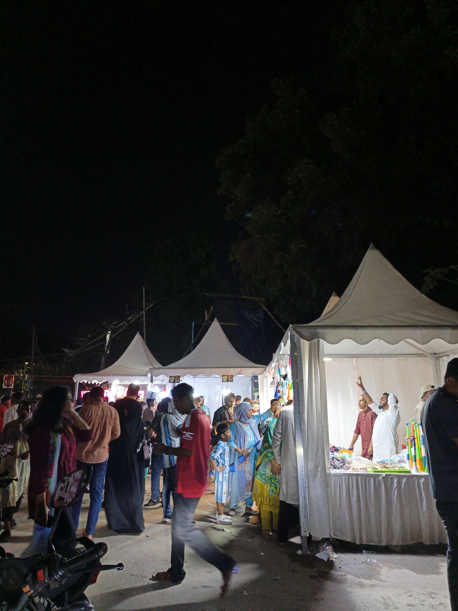 This is a nighttime, medium shot of a street market with several tents. In the foreground, a motorcycle is parked on the left side of the frame. A group of people are walking and standing in front of the tents. The tents are white and peaked. The tent on the right has a table set up with merchandise, and a man in a white shirt is gesturing with his hand. The ground is paved with asphalt, and there is some trash on the ground. The sky is dark, and there are trees visible in the background.