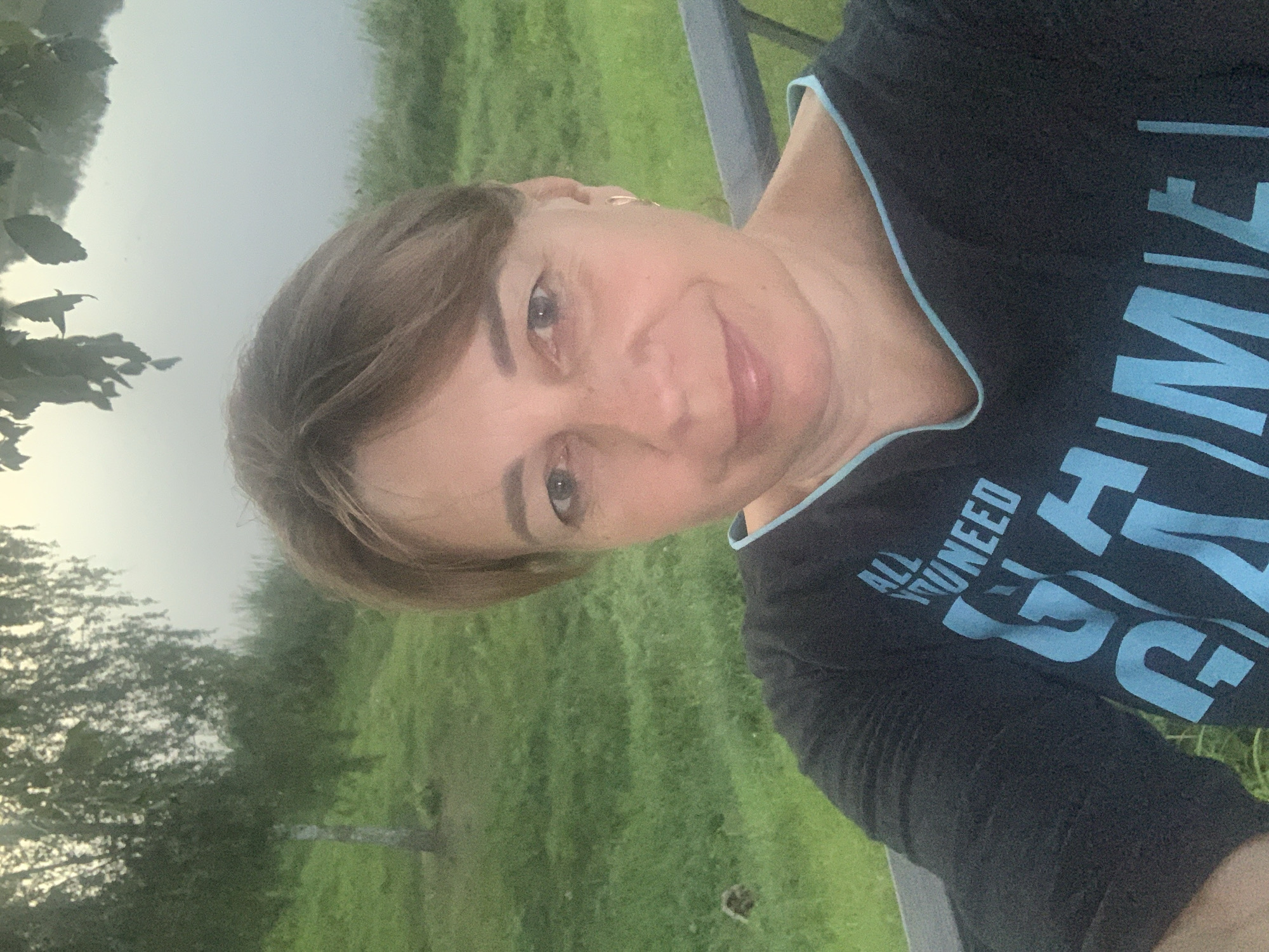 A woman is taking a selfie outdoors. She is looking at the camera with a slight smile. She has shoulder-length brown hair and is wearing a dark t-shirt with light blue lettering that reads "ALL YOU NEED IS GAME". The background is a grassy area with a tree and a body of water. The image appears to have been taken in the city of Kapyĺ, Belarus.