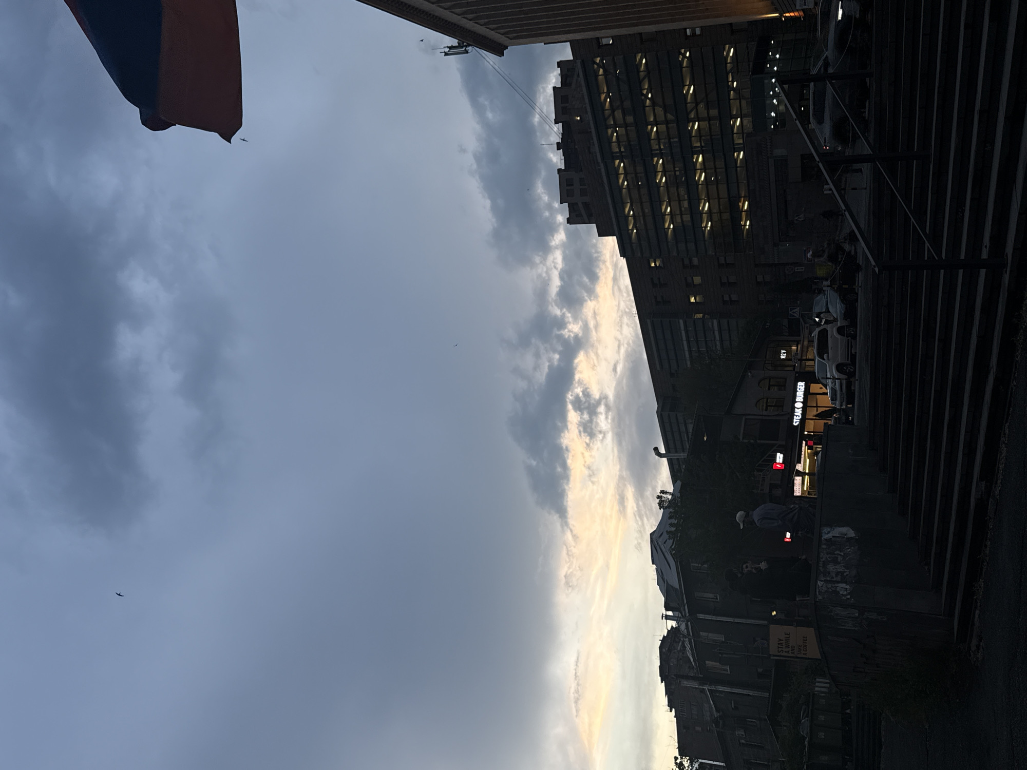 The image shows an evening street scene in Yerevan, Armenia. On the left, a portion of the Armenian flag hangs. The sky is overcast with a line of lighter clouds highlighted by the setting sun. A modern building with lit windows is visible to the right of the lighter clouds. In the foreground, people are visible on a set of stairs and a sign that reads "STAY A WHILE AND TAKE A COFFEE" is visible. The restaurant "Steak Burger" is visible to the right. Cars are parked along the street.