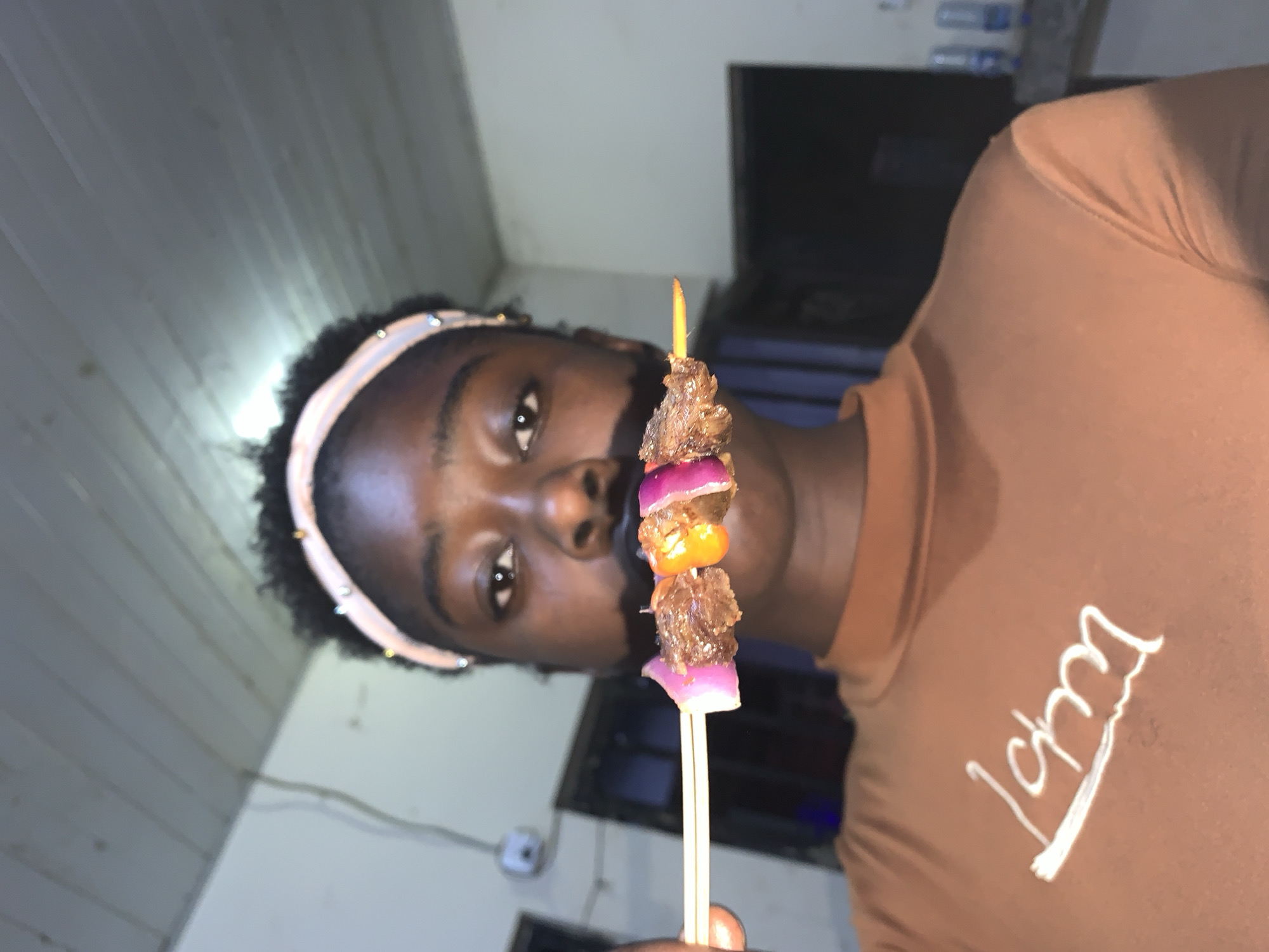 A person with dark skin and short, curly black hair is holding a kebab on a wooden skewer in front of their face. They are wearing a light pink headband and a brown long-sleeved shirt with a white design on the front. The kebab consists of pieces of meat, red onion, and a yellow bell pepper. The background includes a light-colored wall, a dark doorway, and a few unidentified objects. There is no evidence to suggest that the location is in Kabong, Nigeria.