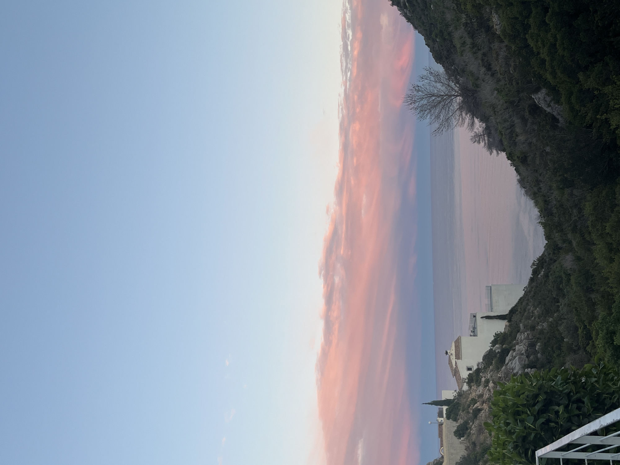 The image depicts a scene in el Poble Nou de Benitatxell/Benitachell, Spain at either sunrise or sunset, oriented in portrait layout. A large, vibrant band of pink-hued clouds stretches vertically across the center of the frame, contrasting with the light blue sky on the left. To the right of the clouds, a rugged cliff covered in green vegetation leads down to the sea. Several white buildings and a church are visible on the slope. In the foreground, a small section of a white metal railing is visible. The ocean is calm and reflects the pink hues of the clouds. A bare tree clings to the cliffside near the sea.