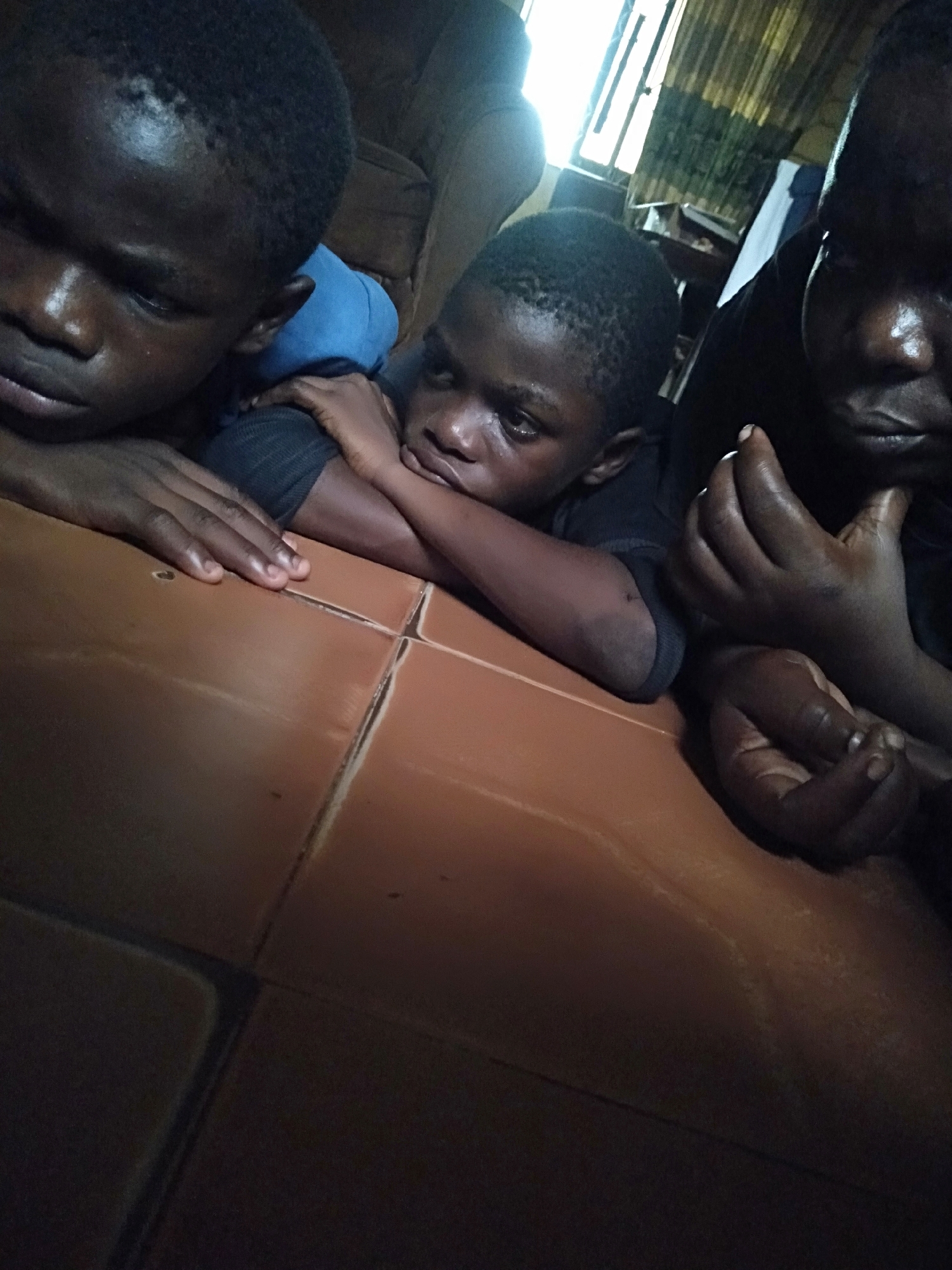 Three children lie prone on a brown tile floor. The child on the left wears a blue shirt and rests their face on their hand. The child in the center wears a dark shirt and looks towards the camera. The child on the right also wears a dark shirt. A window with white curtains is visible in the background. The location is in Shangisha, Nigeria.