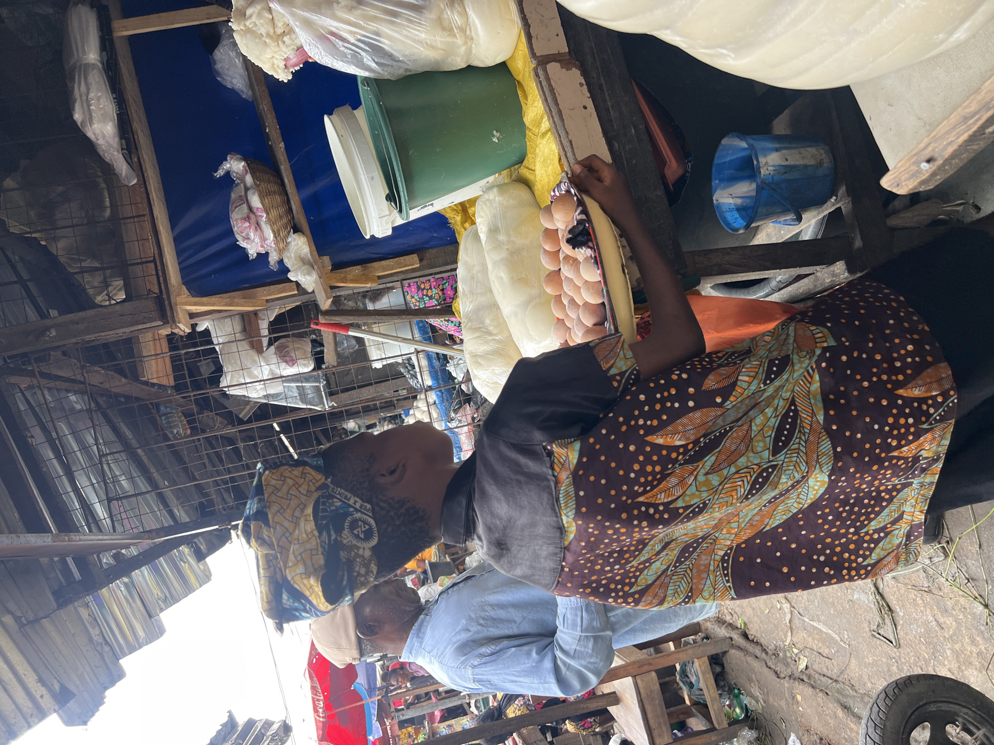 The image shows an outdoor market scene in Nkpor, Nigeria. In the foreground, a person wearing a patterned headwrap and a brown shirt with a colorful leaf and dot pattern is holding a tray of eggs. To the left, a man in a light blue shirt and a tan cap is standing. Behind them, there are market stalls with various goods, including what appears to be food items wrapped in plastic and a basket of items on a shelf. There are also large white sacks or packages on display. The ground is dusty and uneven, and there are wooden structures supporting the market stalls. The overall atmosphere suggests a busy, local marketplace.