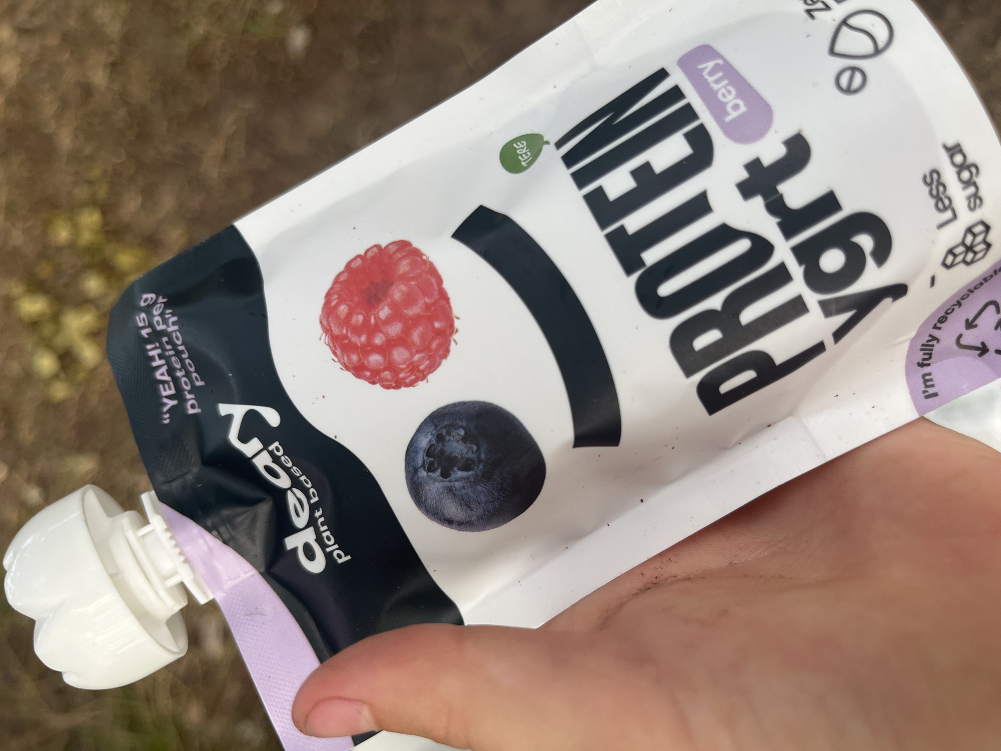 The image shows a close-up of a person holding a pouch of "Deary plant based protein ygrt" in the flavor "berry." The pouch is predominantly white with black and purple text and graphics. A raspberry and a blueberry are depicted on the front of the pouch. The top of the pouch has a white, screw-on cap. The background is blurred and appears to be an outdoor environment with brown and green tones, possibly suggesting a natural setting. There is no visible indication of a location in the city of Pillapalu, Estonia.