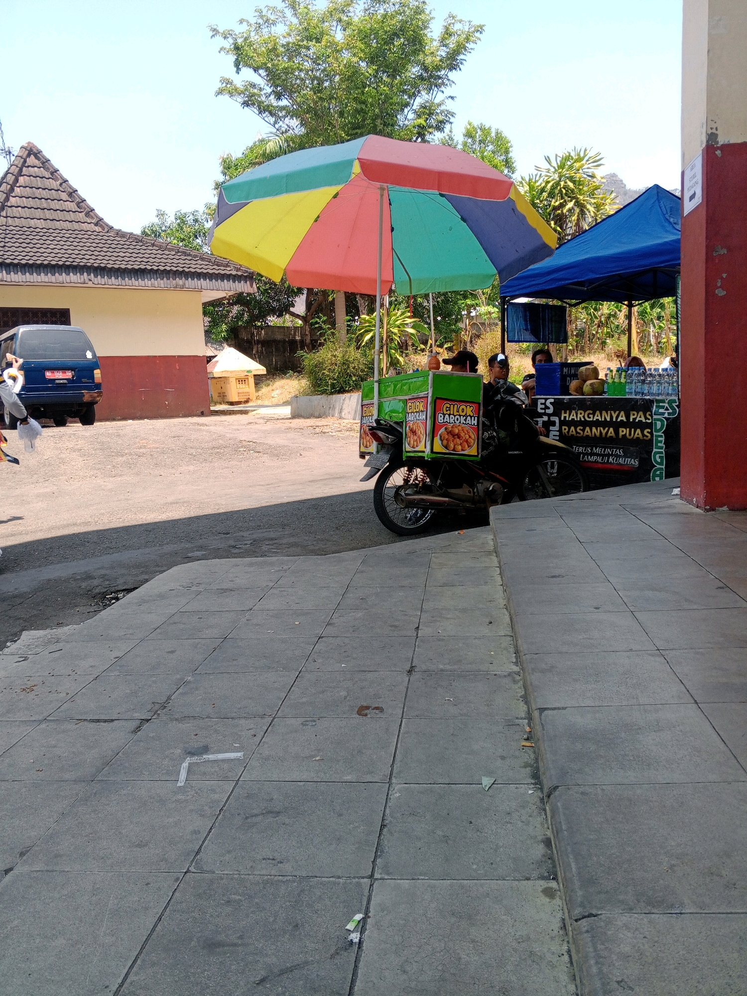 In Wuryorejo, Indonesia, on a bright, sunny day, a street food vendor operates from a mobile cart attached to a motorcycle. The cart is shaded by a colorful, multi-paneled umbrella and is positioned next to a blue tent, likely another vendor's stall. A sign on the food cart advertises "CILOK BAROKAH," and the cart itself displays images of the food being sold. Behind the cart, several individuals are visible, some appearing to be customers and others working at the stalls. The food cart is parked on a paved area, with a tiled sidewalk in the foreground. A blue van is parked nearby to the left, and a building with a tiled roof is visible in the background. The environment suggests a typical street scene in a populated area, possibly a market or a busy roadside.