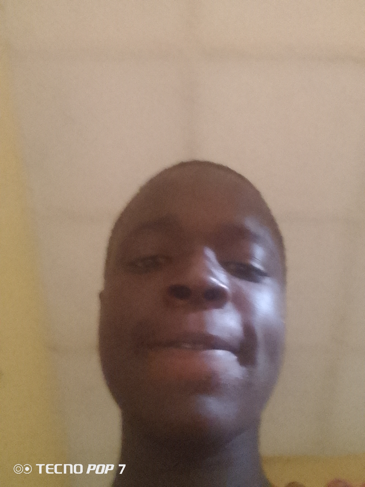 The image is a selfie of a young Black male with short hair, taken from a slightly upward angle. His face is the primary subject, with his skin appearing smooth and slightly reflective. He is looking directly at the camera with a subtle smile, his lips slightly parted. The background is a plain, textured wall, with a gradient of color from light yellow on the left to a beige tone on the right. In the lower left corner of the image, the text "TECNO POP 7" is visible, indicating the device used to take the photograph. The lighting suggests an indoor environment, possibly a home. The scene provides no specific information about the city of Ipetumodu, Nigeria, other than the potential origin of the photograph based on the prompt's context.