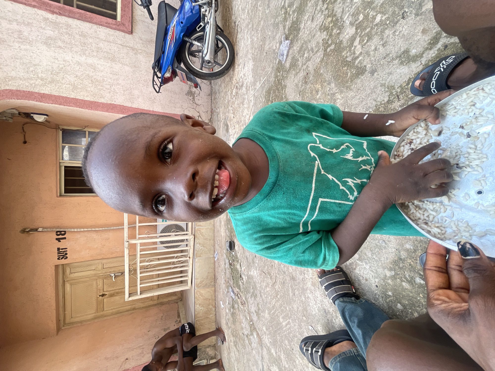 A boy in a green shirt is holding a metal tray filled with rice. He is smiling and sticking his tongue out. There is a motorcycle in the background. A person's legs are also visible, wearing sandals and jeans. A man is seen sitting at the bottom, shirtless. The word "SUIT 18" can be seen on the door. The location is likely Lokoja, Nigeria, given the prompt's instructions.