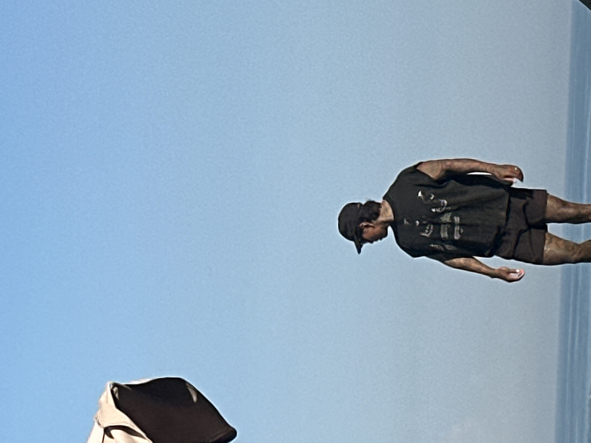 A man wearing a dark sleeveless t-shirt and dark shorts stands on what appears to be a beach in North Kuta, Indonesia. He is looking to his left, with a dark baseball cap on his head. The sky is a clear, pale blue. In the upper left corner of the image, a portion of a light-colored umbrella is visible. The ocean is visible in the background, with a calm horizon.