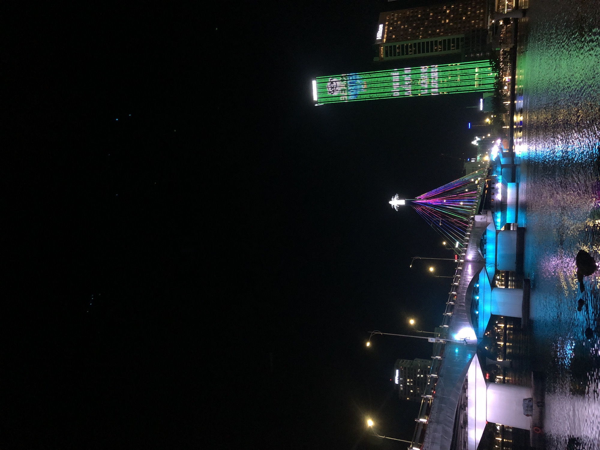This is a nighttime, eye-level photograph of the Dragon Bridge in Đà Nẵng, Vietnam, spanning the Hàn River. The bridge is illuminated with colorful lights that reflect on the water. The bridge's structure features a large, white arch with cables supporting the road deck, which is also lit with blue and white lights. A statue of a figure with outstretched arms, adorned with multicolored lights, stands atop the bridge's apex.
To the right of the bridge, a tall, slender skyscraper is illuminated with bright green vertical lights, forming a patterned design that resembles a character or image. Above this skyscraper, a smaller building displays the "Hilton" logo. The dark sky above is largely devoid of stars, with only a few faint specks visible. The river in the foreground is dark and reflects the lights from the bridge and the buildings. The overall scene depicts a vibrant urban nighttime landscape.