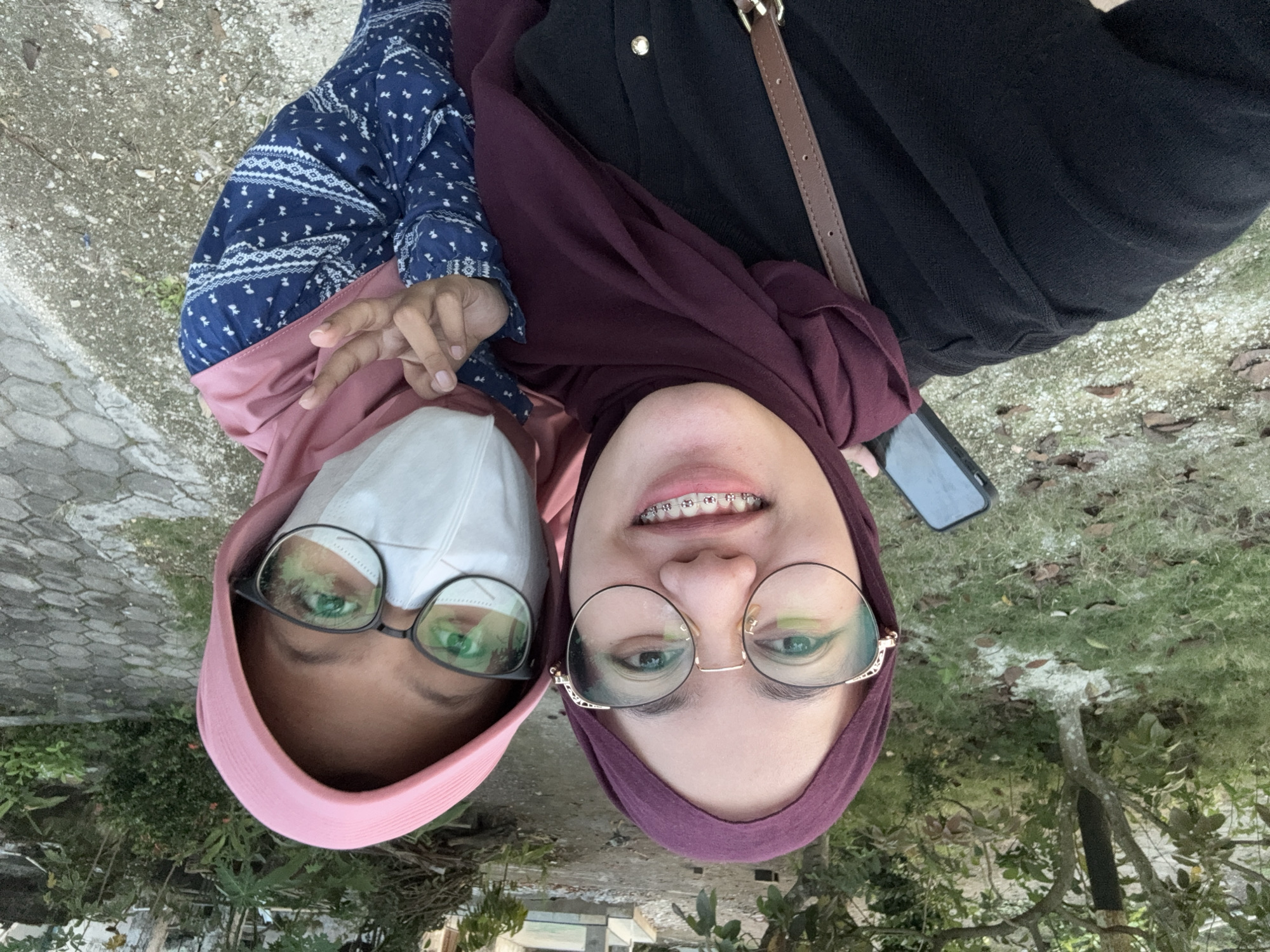 Two women are positioned closely together outdoors. The woman on the left wears a light pink head covering and a white face mask, along with black-framed glasses. She is also wearing a patterned blue long-sleeved shirt. The woman on the right is wearing a maroon head covering and round, wire-rimmed glasses. She is also wearing a black top with a brown purse strap over her right shoulder and has braces on her teeth. Her phone is visible in the top right.
The location appears to be an outdoor area in Gadingrejo, Indonesia, with greenery and a paved surface in the background. The photo is taken from a low angle, tilting upwards, and the horizon is not visible.