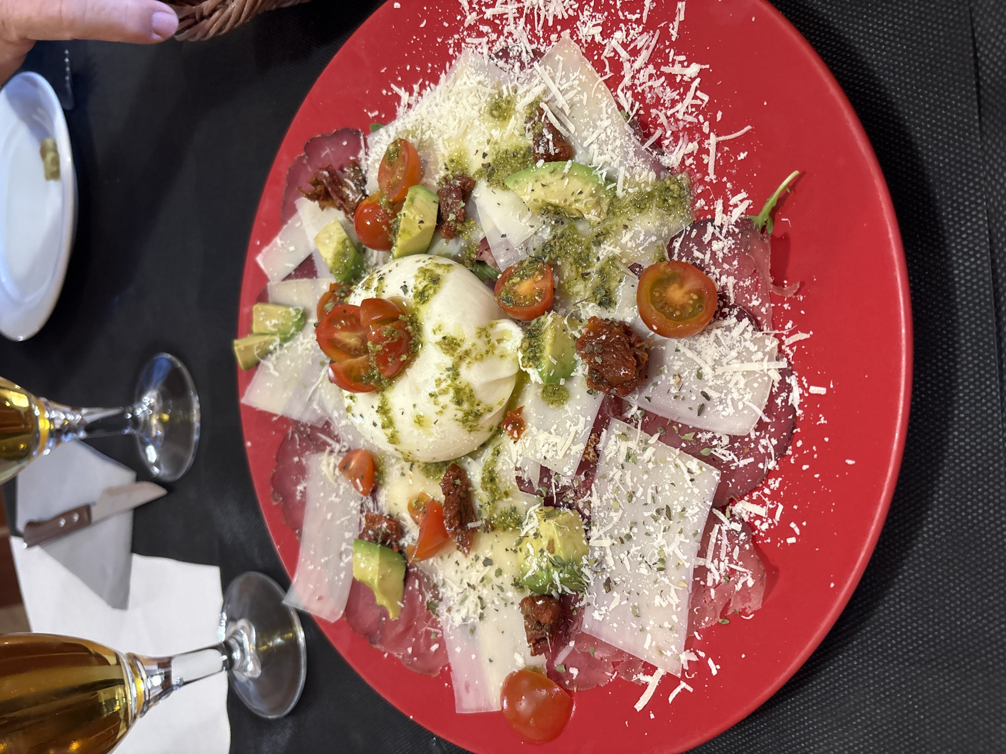 The image shows a red plate of food, likely a salad or appetizer, with a central ball of burrata cheese, cherry tomatoes, avocado, sun-dried tomatoes, and shavings of cheese on top of thinly sliced meat. The plate is on a table covered with a black surface. There are two wine glasses with a light-colored liquid, possibly white wine, visible in the lower left corner of the image. A knife and a white plate are near the glasses. Part of a person's hand is visible in the top left corner. The location is likely a restaurant in Teulada, Spain, based on visual cues (food style). The scene appears to be set during a meal.