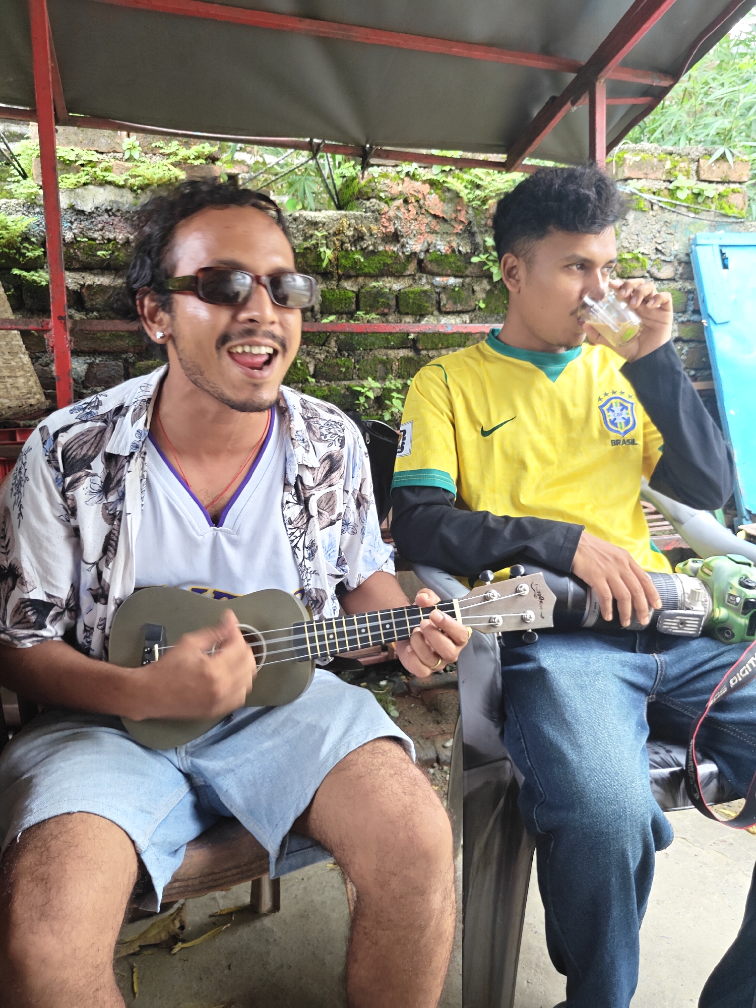 Two men are seated outdoors under a covered structure, possibly in Pokhara, Nepal, with a brick wall covered in moss and vegetation behind them. The man on the left, wearing sunglasses and a floral print shirt over a white athletic shirt, is playing a ukulele and appears to be singing. He is wearing light blue denim shorts. The man on the right is wearing a yellow and green Brazil soccer jersey with black long sleeves underneath. He is holding a glass in his right hand and drinking from it. He has a large camera lens resting on his lap. The scene appears to be casual and relaxed.