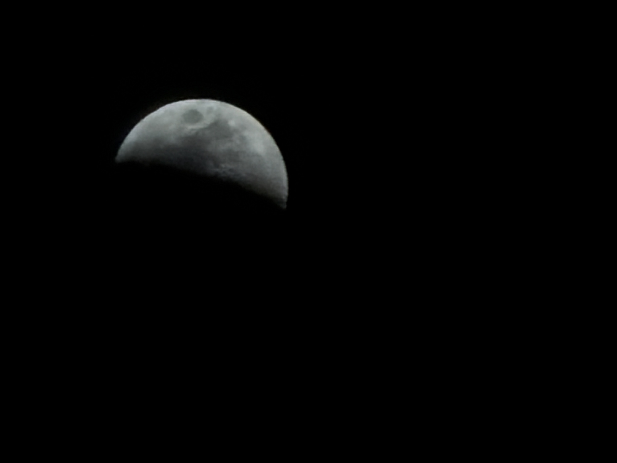 The image shows a waxing gibbous moon in the upper left quadrant against a completely black background. No information is available to determine if this image was taken in Izhevsk, Russia. There are no discernible objects, people, actions or interactions visible in the frame besides the moon. The environment is dark.