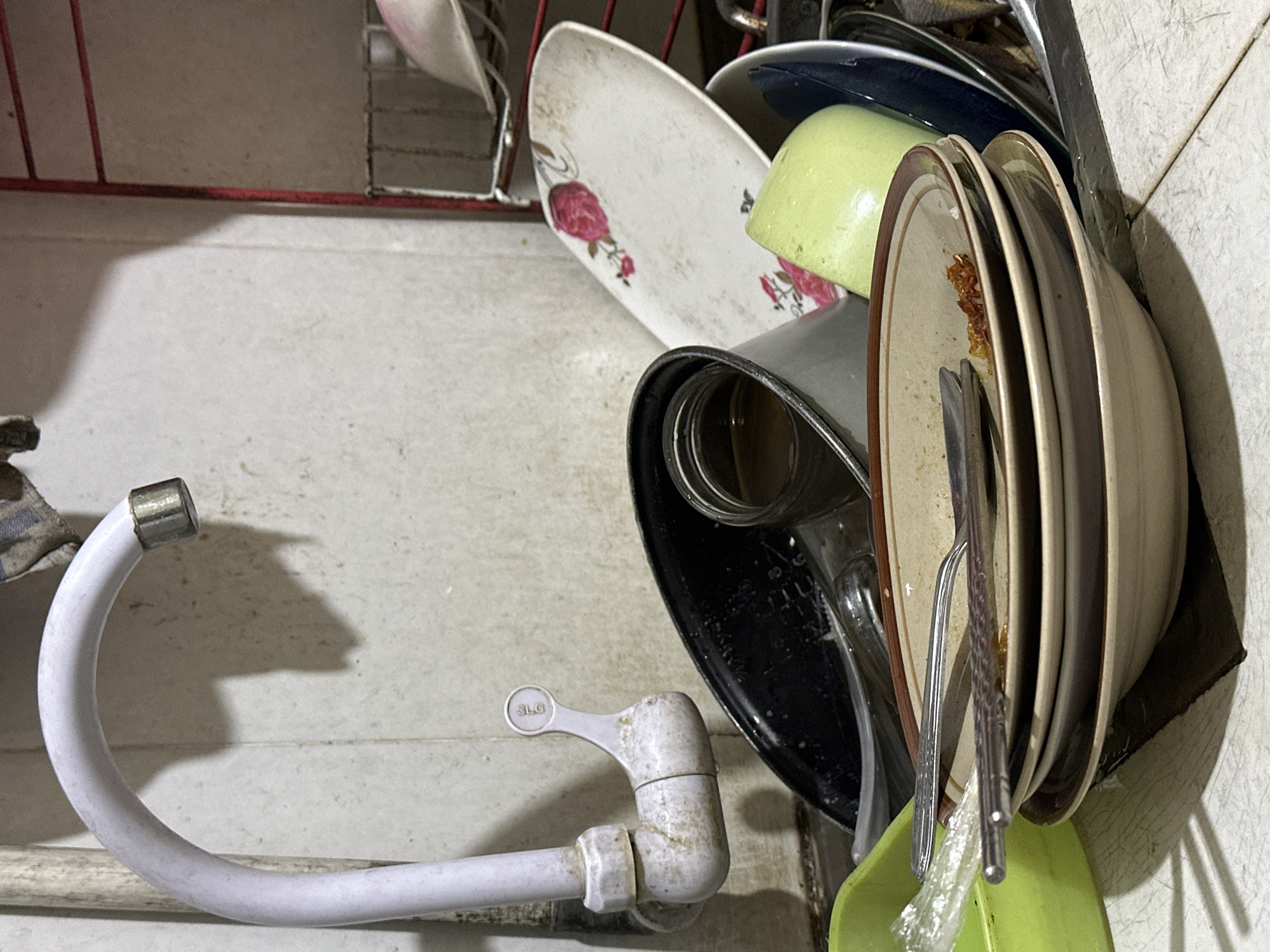 A close-up, eye-level shot captures a sink filled with unwashed dishes. In the upper left, a white faucet with a chrome tip is visible, casting a shadow on the tiled wall behind it. The faucet is dirty and appears to be made of plastic. To the right of the faucet, a rack with red metal bars holds various items, including a pink-capped object.
The sink itself is overflowing with dirty plates, bowls, and a glass jar containing a dark liquid. A bright green plastic basin is partially visible on the left. A stack of three beige plates with brown rims is prominent in the foreground, with a fork and spoon resting on the top plate. Another large, white oval plate with a floral pattern of pink roses is leaning against the dishes. A pale green bowl or cup is also visible amongst the clutter. The overall impression is one of domestic disarray in a kitchen or utility area. The scene is likely located in a residential setting in Bulakrejo, Indonesia, given the context provided.