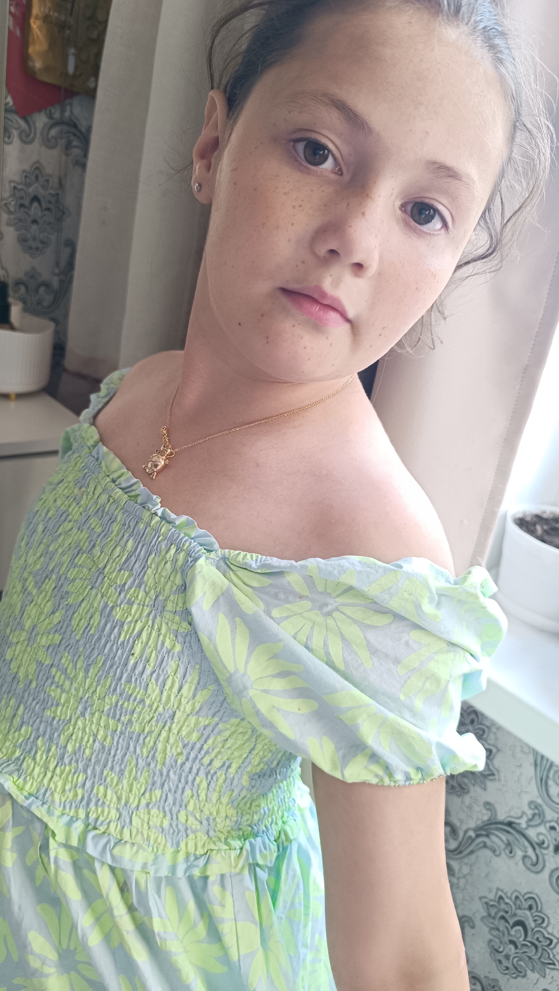 A young Caucasian girl with brown hair and brown eyes is pictured indoors, with a soft focus on the background. She has a dusting of freckles across her nose and cheeks. She is wearing a light blue and green floral print dress that is off-the-shoulder. A delicate gold necklace with a small pendant is visible around her neck. The girl's gaze is directed slightly away from the camera. The environment appears to be a domestic setting, with hints of curtains and patterned wallpaper visible. There is a plant in a white pot on a windowsill to the right of the frame. The image is taken in Chaikovsky, Russia.