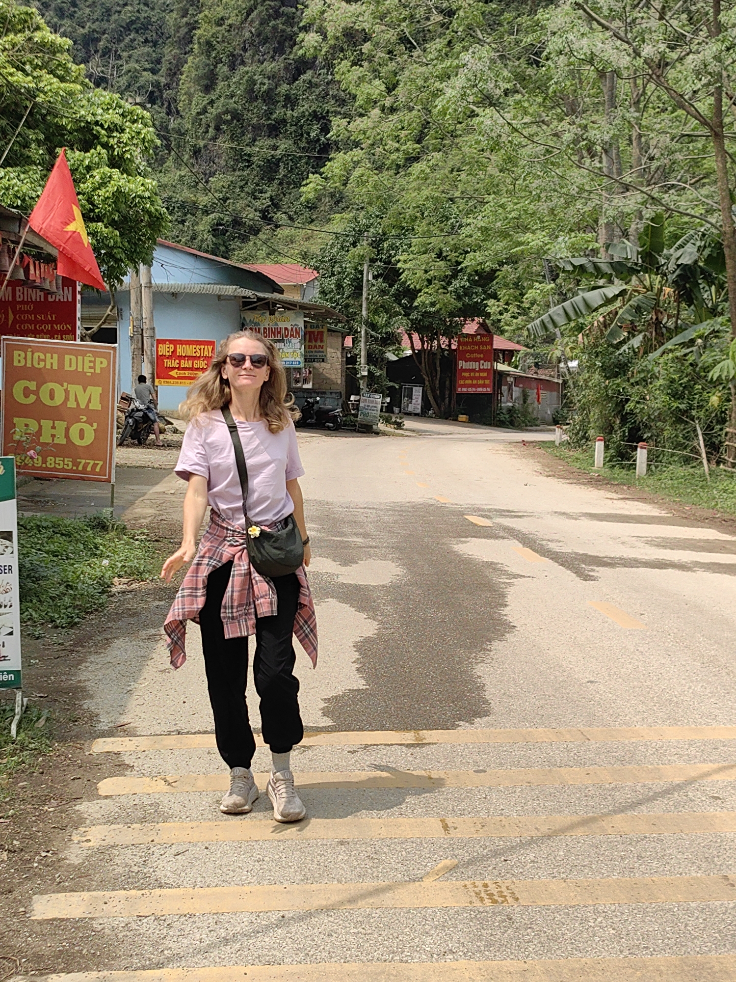 A woman wearing sunglasses, a lilac t-shirt, black pants, and grey sneakers stands in Xã Đàm Thủy, Vietnam. A pink and black plaid shirt is tied around her waist, and she wears a black bag over her shoulder. She stands on a road marked with yellow zebra stripes, with a faded yellow center line. Signs in Vietnamese are on either side of the road, and a red Vietnamese flag is visible on the left. Lush green foliage and a mountain rise in the background. The sky is overcast.