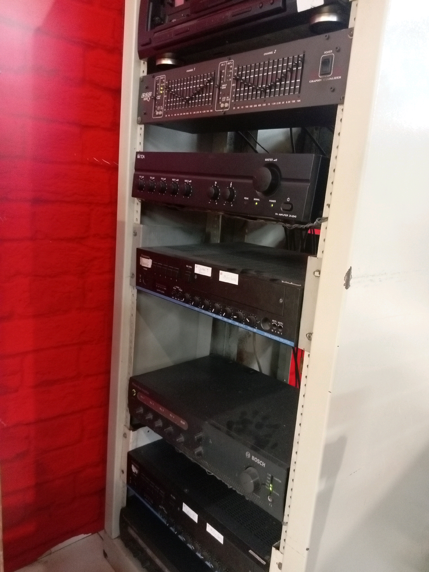 The image shows a rack of audio equipment, possibly in a sound studio or performance venue. The rack contains several black audio devices, including graphic equalizers, amplifiers, and mixers. The devices are stacked vertically, with some wires visible between them. The rack itself is a light-colored metal structure. The background on the left is a textured red wall, and the background on the right is a plain white wall. The overall lighting is neutral, suggesting an indoor setting. There are no people visible in the image.
This image does not contain enough information to determine the specific location in Batam, Indonesia.