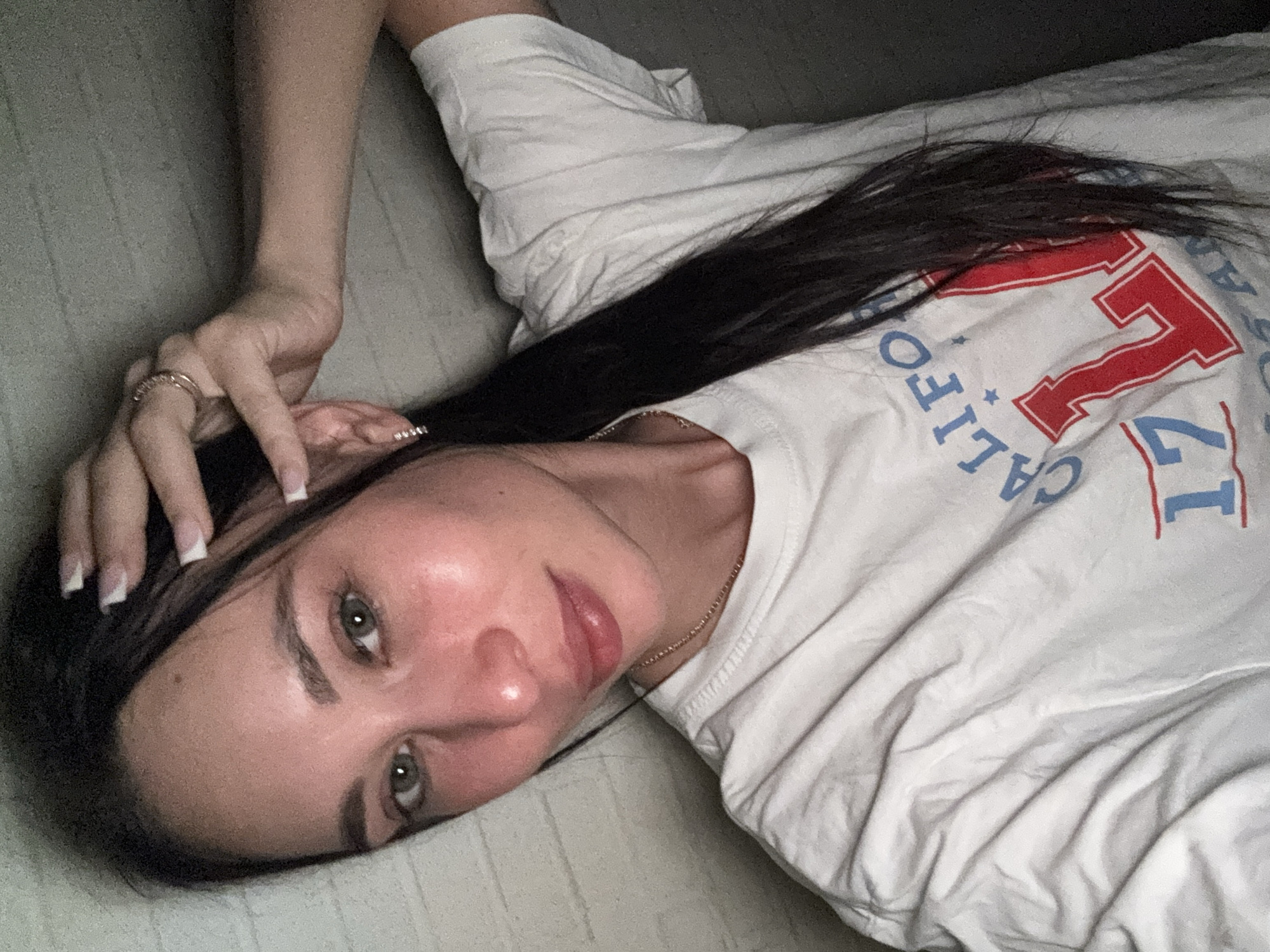 A young woman with dark hair and light eyes is lying on a light-colored surface. She is wearing a white t-shirt with "California 77" printed on it in blue and red lettering. Her left hand is raised near her head, adorned with rings. A thin gold chain is visible around her neck. The background appears to be a plain, light-colored surface, possibly a bed or sofa. The image's location in Chaikovsky, Russia, is not verifiable from the image alone.