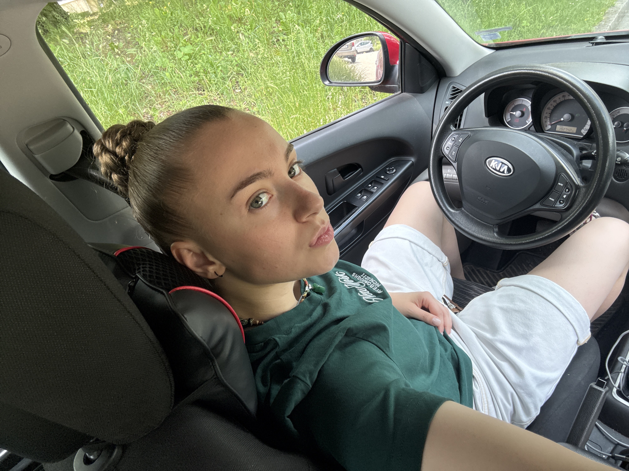 A young woman with a braided bun hairstyle is seated in the driver's seat of a car, looking at the camera. She is wearing a dark green t-shirt and white shorts. The car's interior is visible, including the steering wheel with a Kia logo, the dashboard with gauges, and the door panel with window controls. Outside the car, green grass and trees are visible, suggesting an outdoor setting. The sunlight indicates it is daytime, and the weather appears to be fair. A side mirror reflects two other vehicles. The woman's expression is neutral, and the overall mood of the image is casual and relaxed. The visible text on the t-shirt is "FIVE BOROUGHS MIDNIGHT".