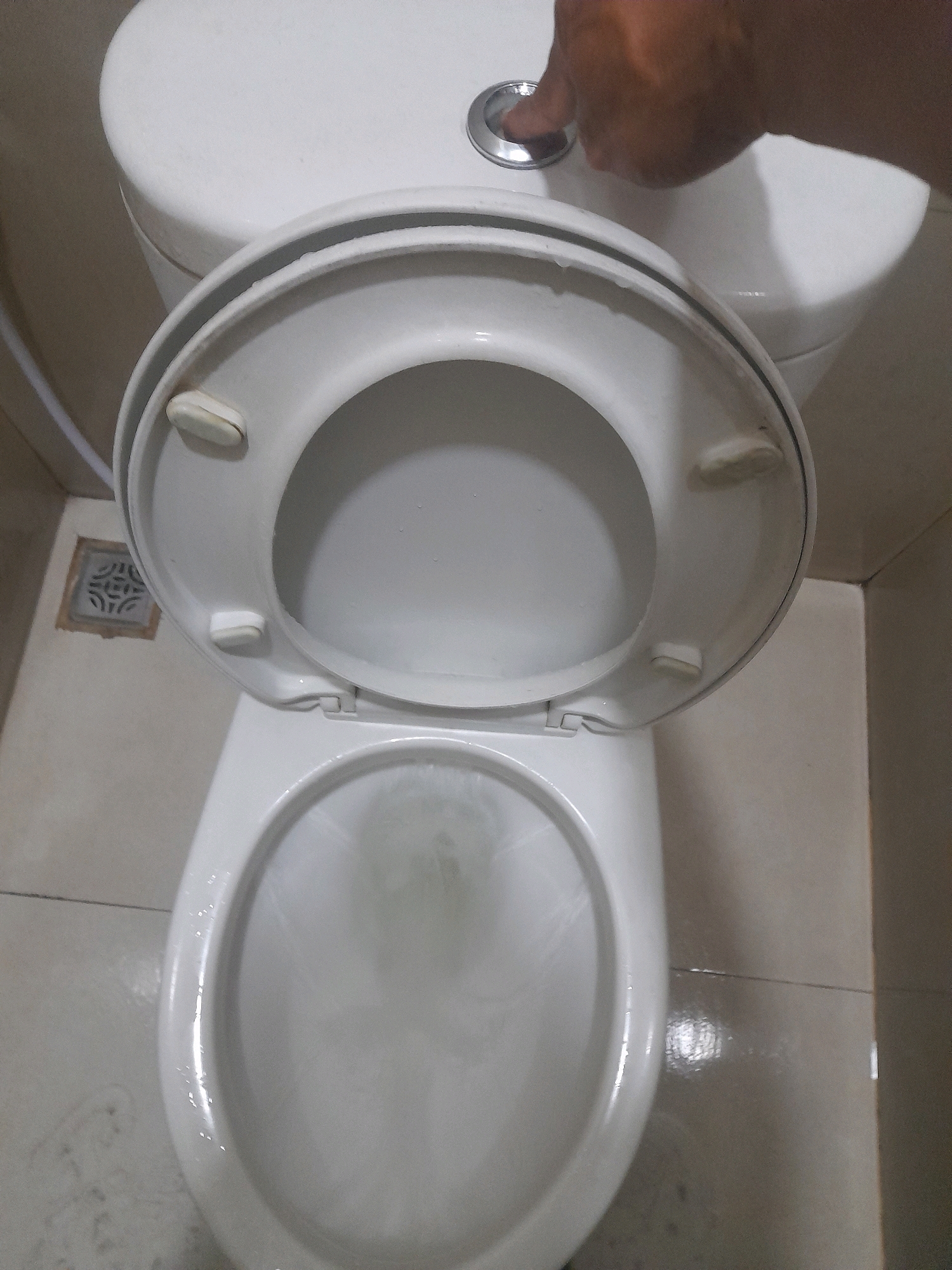 The image shows a close-up, overhead view of a white toilet with the lid and seat up. A hand is pressing the flush button located on top of the toilet tank. The toilet bowl contains water, and there are water droplets visible on the toilet seat. The surrounding environment appears to be a tiled bathroom with beige walls. A drain grate is visible on the floor to the left of the toilet. The scene is consistent with a bathroom in Surabaya, Indonesia.