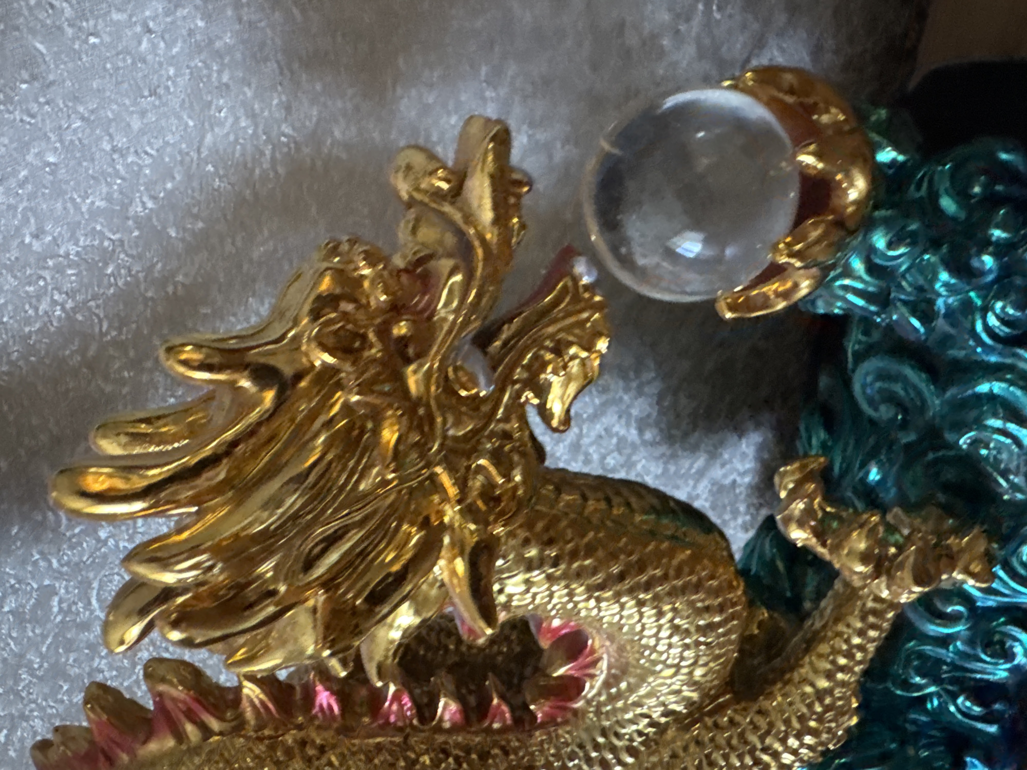 The image shows a close-up of a golden dragon figurine with intricate scales and a decorative mane. The dragon is clutching a clear crystal ball in its right claw, which is supported by a gold-colored, multi-fingered base. The figurine appears to be resting on a base of swirling, metallic teal waves. The background is a textured, light-colored surface. There is no indication of the city of Svitlovodsk, Ukraine, or any other specific location. The image focuses solely on the detailed craftsmanship of the dragon figurine and its accessories.