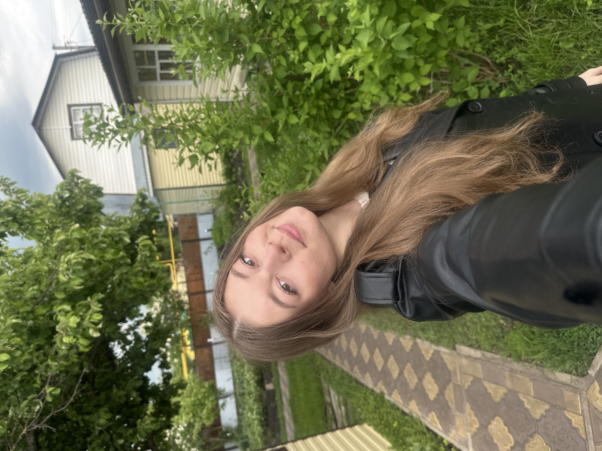 A young woman with long brown hair wearing a black leather jacket is taking a selfie outdoors. The setting appears to be a residential area with a house visible in the background, surrounded by green trees and grass. A paved walkway is also visible. The location is identified as Коноваловка, Russia.