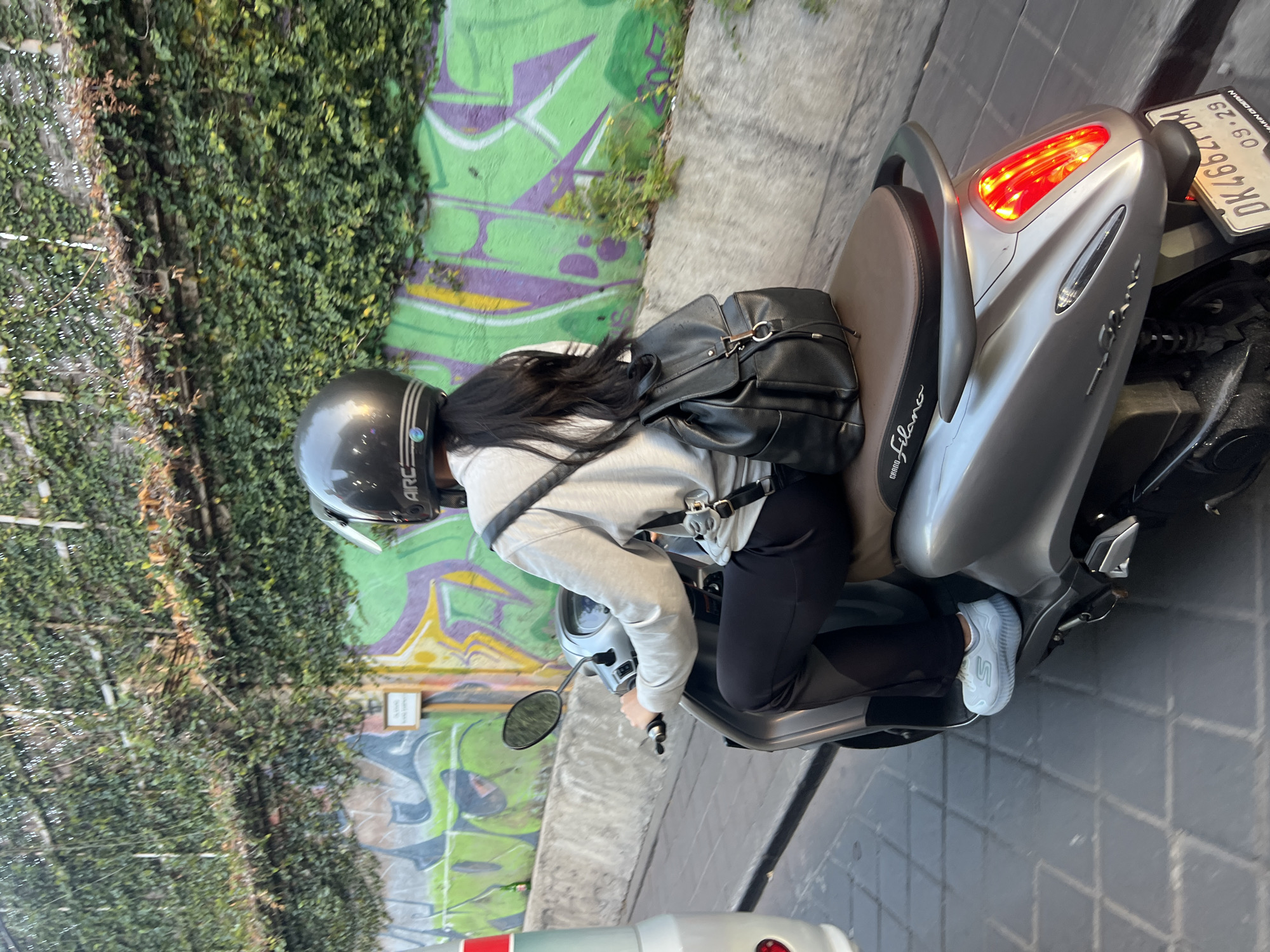 A person wearing a gray hoodie, black pants, and a dark gray helmet with a silver stripe is riding a silver scooter. They are carrying a black backpack. The scooter has a brown seat with "GRAND FILANO" printed on it. The license plate reads "DK 4662DUM" and "09-29". The scooter is on a brick-paved street. Behind the scooter is a concrete wall covered in green ivy and graffiti. The location is Kuta, Indonesia.