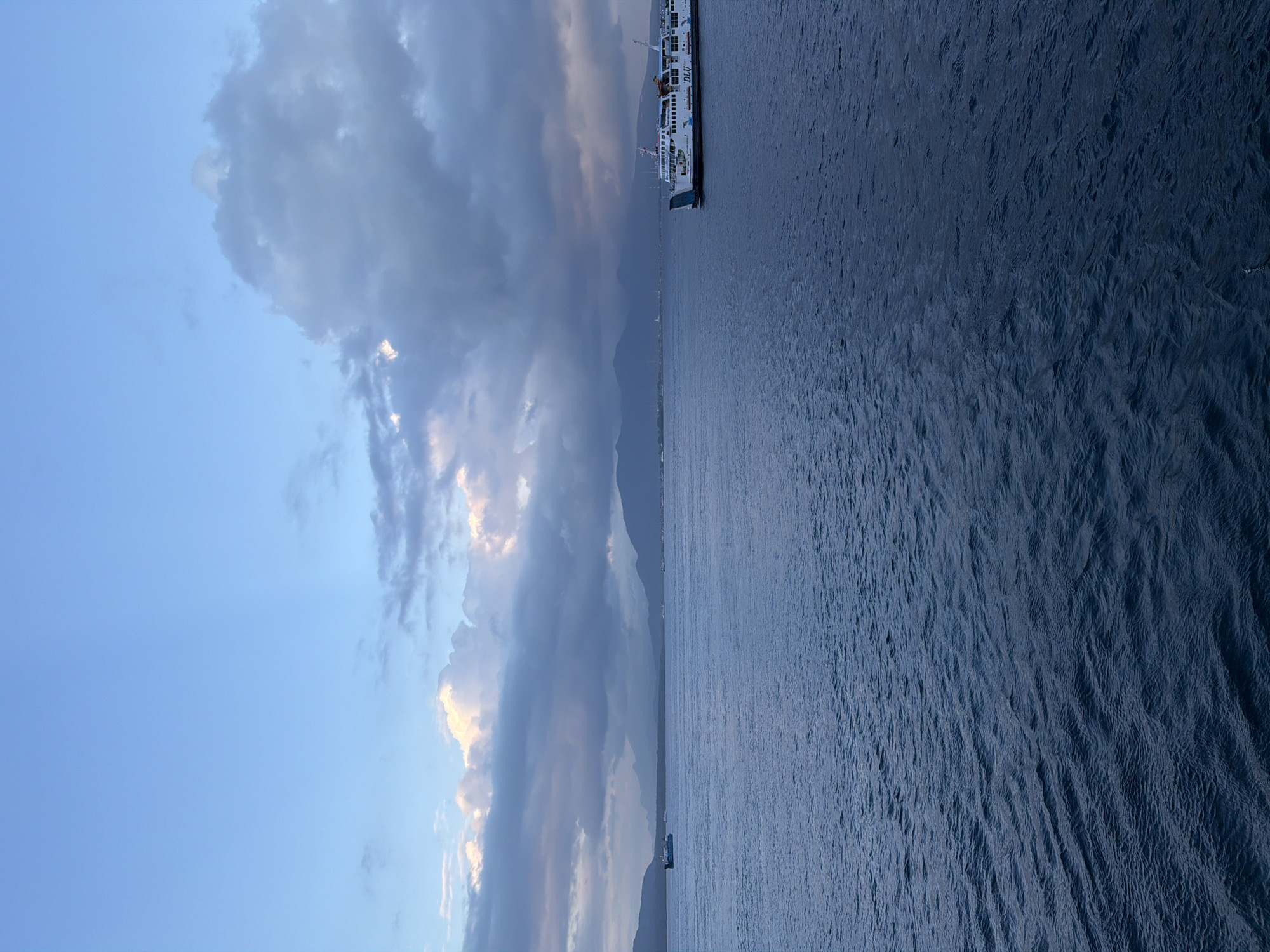 A wide-angle, medium-shot photograph shows a serene seascape in Ketapang, Indonesia, at dusk. The sky is a gradient of pale blue to darker hues, with dramatic, textured clouds in shades of gray and white. Sunlight breaks through the clouds in places, casting a warm glow. In the distance, a range of hazy mountains forms the horizon. The foreground is dominated by a vast expanse of dark blue water, with gentle ripples reflecting the twilight sky. Two ferries are visible on the water. A smaller, blue and white ferry is positioned on the left side of the image, moving away from the viewer. On the right side, a larger, white ferry with blue accents, identified by the name "DLU" on its side, travels towards the right. The overall atmosphere is calm and tranquil, indicative of the transition from day to night.