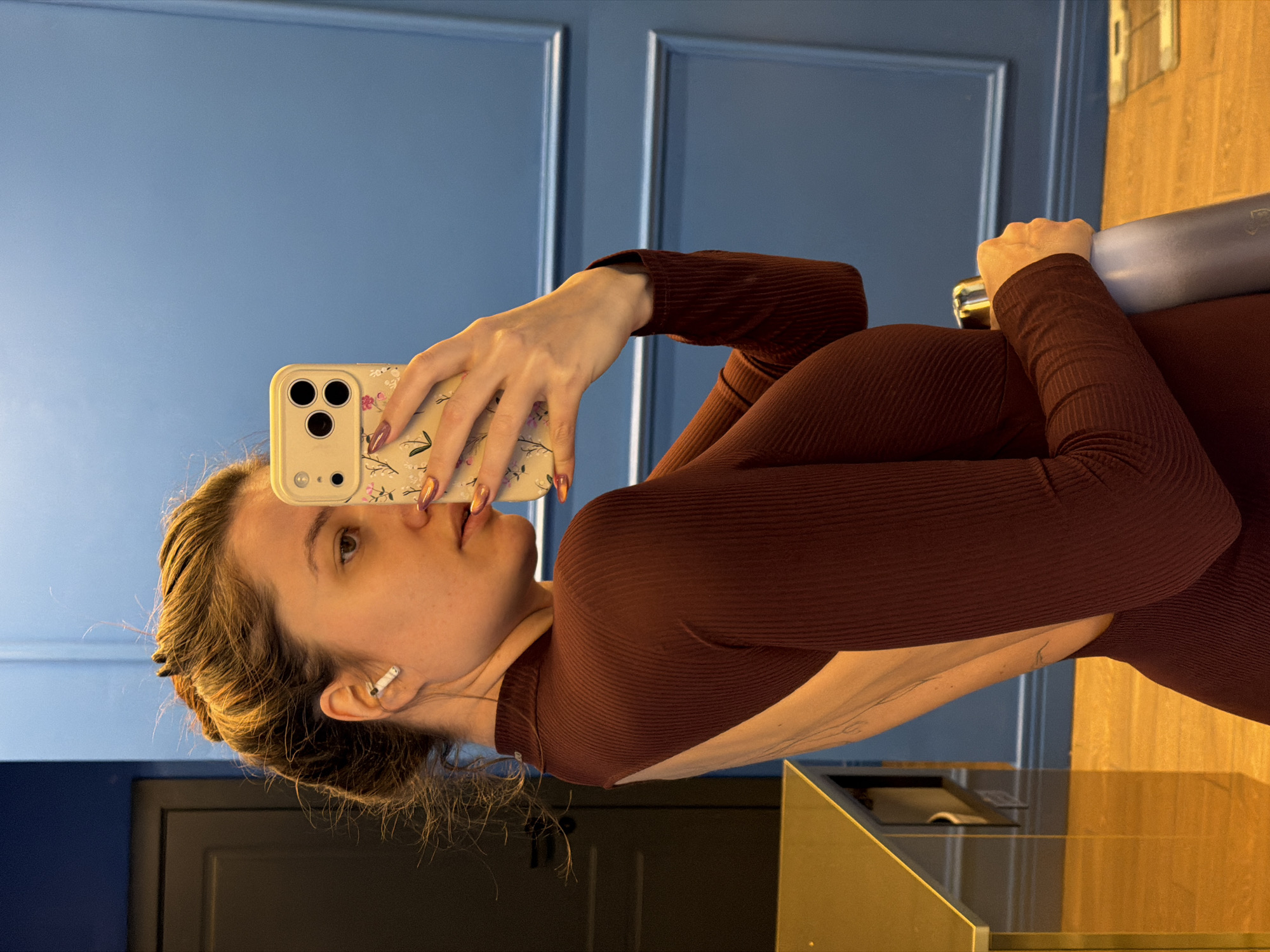 The image is a mirror selfie, rotated 90 degrees clockwise, showing a young woman indoors. She has light skin and brown hair styled back. She is wearing a long-sleeved, ribbed, dark red or burgundy top with an open back. A white earbud is visible in her left ear.
The woman is holding a smartphone with her left hand, facing the camera to take the photo. The phone appears to be an iPhone model with a triple camera setup, housed in a light-colored case adorned with a delicate floral pattern. Her fingernails are long and painted with a metallic, iridescent polish that shifts between orange, gold, and pink hues. A faint, intricate tattoo is visible on her left arm or shoulder blade, exposed by the open back of her top. Her right hand, partially visible, is grasping a silver or grey cylindrical object, possibly a water bottle, behind her. A mirrored and partially obscured logo and text are visible on this object.
The background features an interior space with prominent blue walls that have decorative molding. To the right, there are elements of golden or wooden paneling, suggesting a polished environment. In the lower left, a dark, possibly framed door or a reflection of one is visible. The scene appears brightly lit, indicating either daytime or well-lit artificial lighting. The activity is the woman taking a mirror selfie. The location is an indoor setting within Belgrade, Serbia. No explicit text is clearly legible apart from the ambiguous text on the bottle.