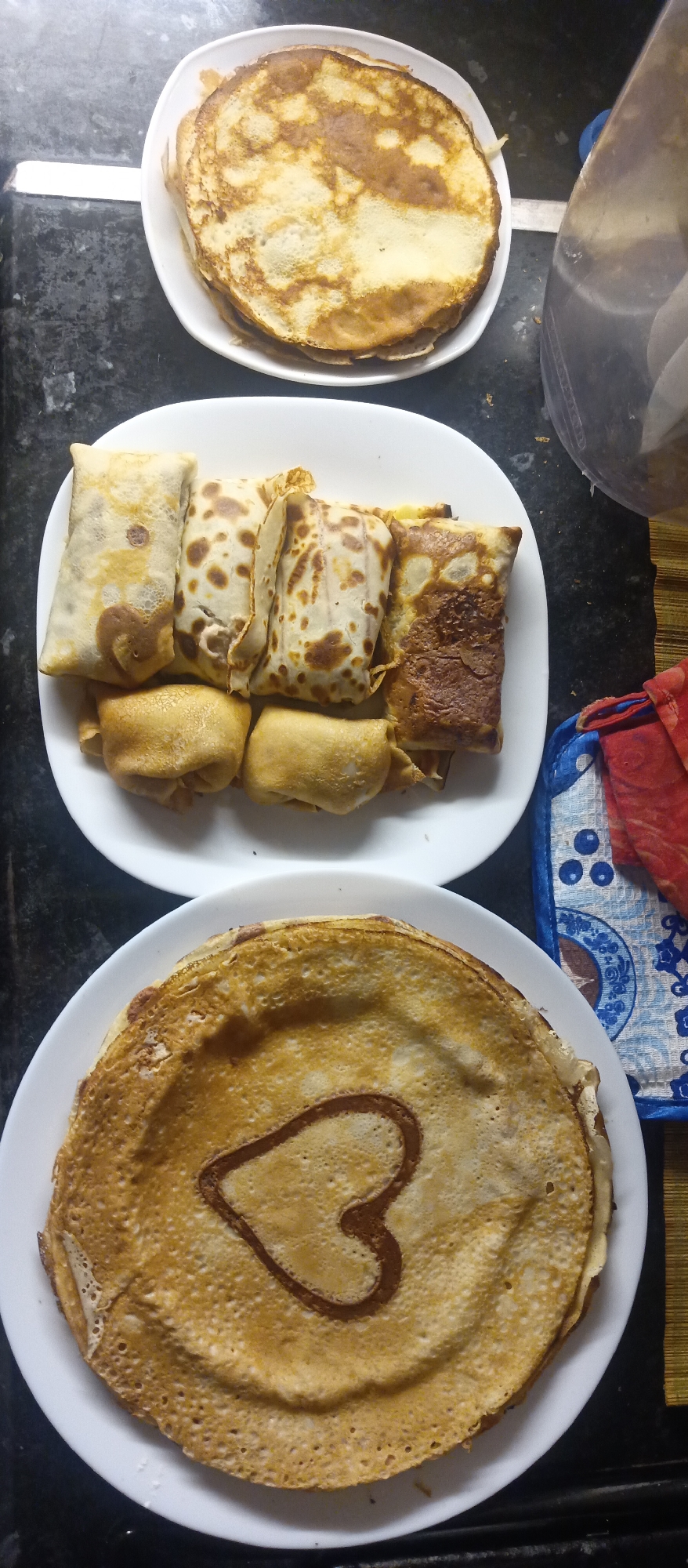 The image shows three white plates of pancakes on a black marble countertop. The top plate contains a stack of round, flat pancakes. The middle plate contains what appear to be filled and rolled pancakes, with some rolled more tightly than others. The bottom plate contains a stack of pancakes, with the top one decorated with a chocolate heart. A red and blue patterned potholder and a kitchen utensil are visible near the plates. The location appears to be a kitchen.
