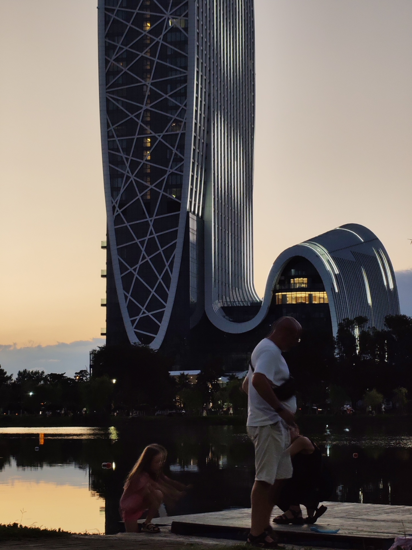 This is a photograph taken at dusk, possibly from a waterfront boardwalk, showcasing a striking modern architectural complex on the left. The complex features two main buildings: a tall, slender skyscraper with a distinctive white geometric lattice pattern against its dark facade, and a lower, curved building with illuminated horizontal lines. The sky is a soft gradient of pale orange and beige, transitioning to a darker dusk sky above.
In the foreground, on a wooden deck or pier, are three people. A young girl, wearing a pink dress and shorts, is crouched down, seemingly interacting with something near the water's edge. To her right, a man in a white t-shirt and light-colored shorts stands, with a dark bag slung across his chest. He appears to be interacting with a second person, who is mostly obscured from view, kneeling or squatting in front of him.
Behind the people and the pier, a body of water reflects the dusky sky and the lights from the distant buildings and streetlights. Trees line the far bank of the water, silhouetted against the twilight. The overall scene suggests a relaxed evening ambiance in an urban waterfront setting.
The architectural style and the specific design of the buildings, particularly the prominent skyscraper with its lattice facade and the curved, illuminated building, are characteristic of the modern development in Batumi, Georgia, especially along its coastline. While the exact location is not explicitly stated, this architectural pairing strongly suggests a view of the Batumi Boulevard area or a nearby waterfront development in the city.