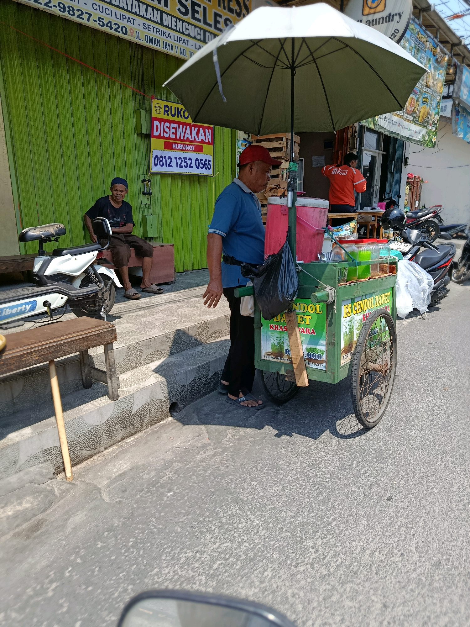 In Bekasi, Indonesia, a street scene unfolds on a sunny day. A man in a blue shirt and red hat stands beside a green food cart selling "Es Cendol Dawet." The cart is filled with various drinks and containers. To the left, an older man in a black t-shirt and dark pants sits on a wooden bench near a white electric scooter. Behind them, a green corrugated metal shuttered shop is visible, with a sign that reads "RUko DISEWAKAN" (Shop for Rent) and a phone number. Further down the street, a man in an orange jacket, possibly a food delivery worker, is seen near another shop. Motorcycles are parked in the background. The scene is set on a paved street with a textured sidewalk.