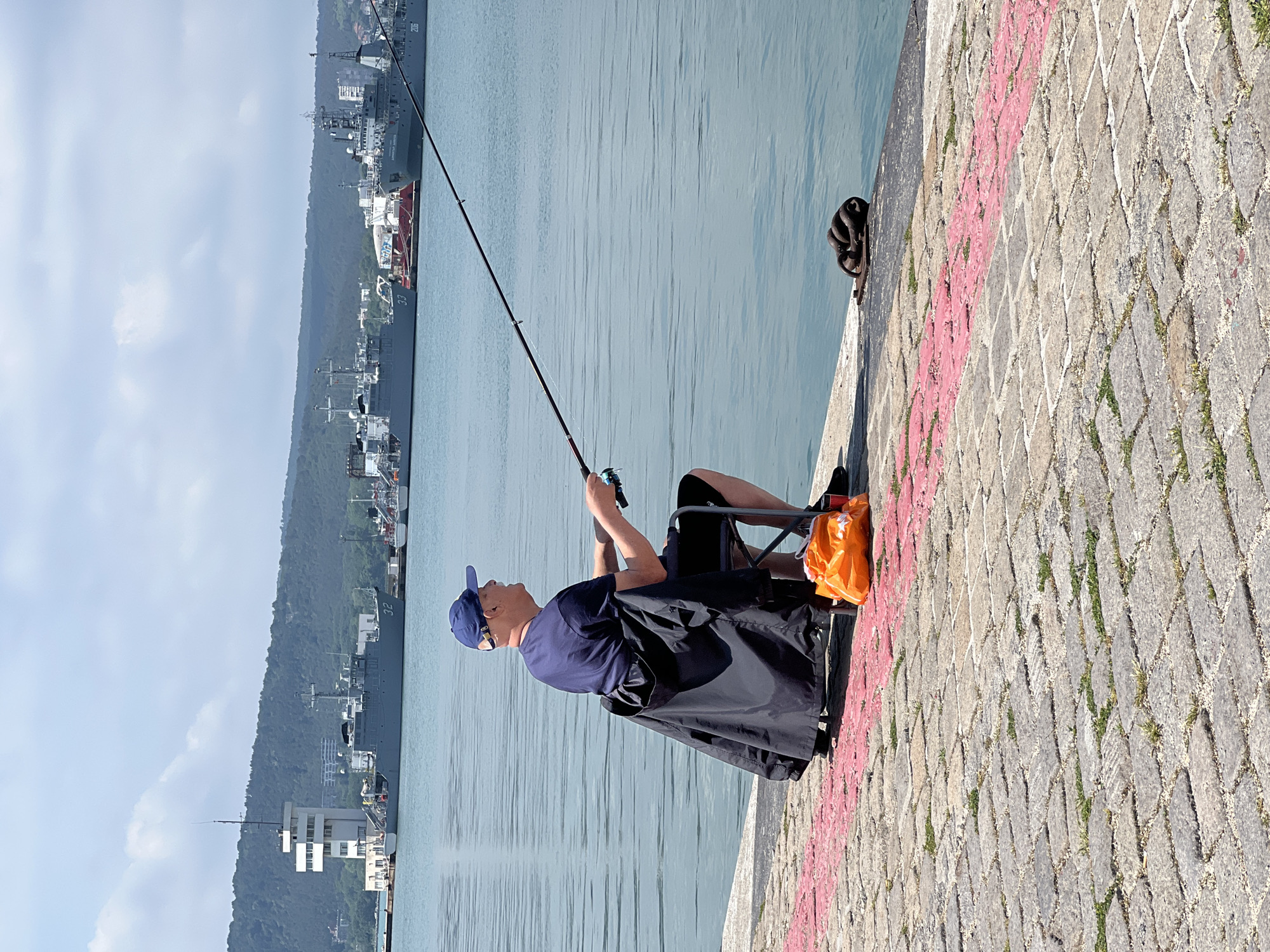 The image, taken in Varna, Bulgaria, depicts a man fishing from a paved pier on a bright, clear day. The man is seated on a black folding chair, wearing a dark blue t-shirt, dark shorts, a blue baseball cap, and sunglasses. He holds a fishing rod, casting its line into the calm, light blue water of the harbor. An orange plastic bag is placed next to his chair on the pavement.
The pier surface is made of grey, interlocking paving stones, with a weathered red painted line running along a section of it. A dark metal bollard or mooring cleat is visible at the water's edge.
In the background, several large ships are docked, their grey hulls suggesting they might be naval or commercial vessels. The numbers "37" and "206" are faintly visible on the sides of some of these ships. Beyond the ships and the water, a prominent, tree-covered mountain or hilly coastline rises under a sky with scattered white clouds. The scene conveys a sense of peaceful recreation set against an industrial maritime backdrop.