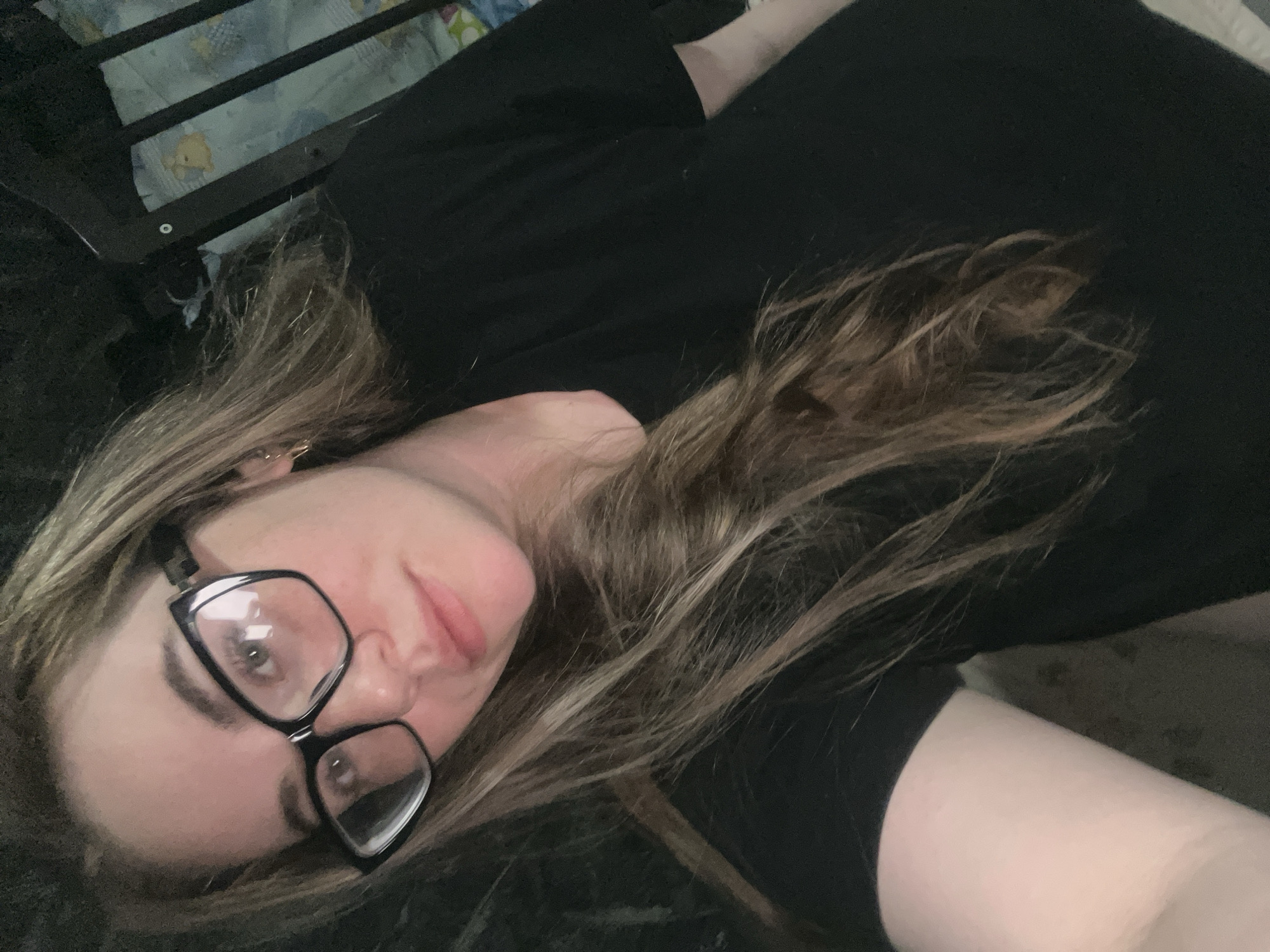 A woman with light brown hair and black-framed glasses is lying down. She is wearing a black shirt. There is a crib with a patterned lining in the background. It is not possible to ascertain whether the location is Chaikovsky, Russia from this image.