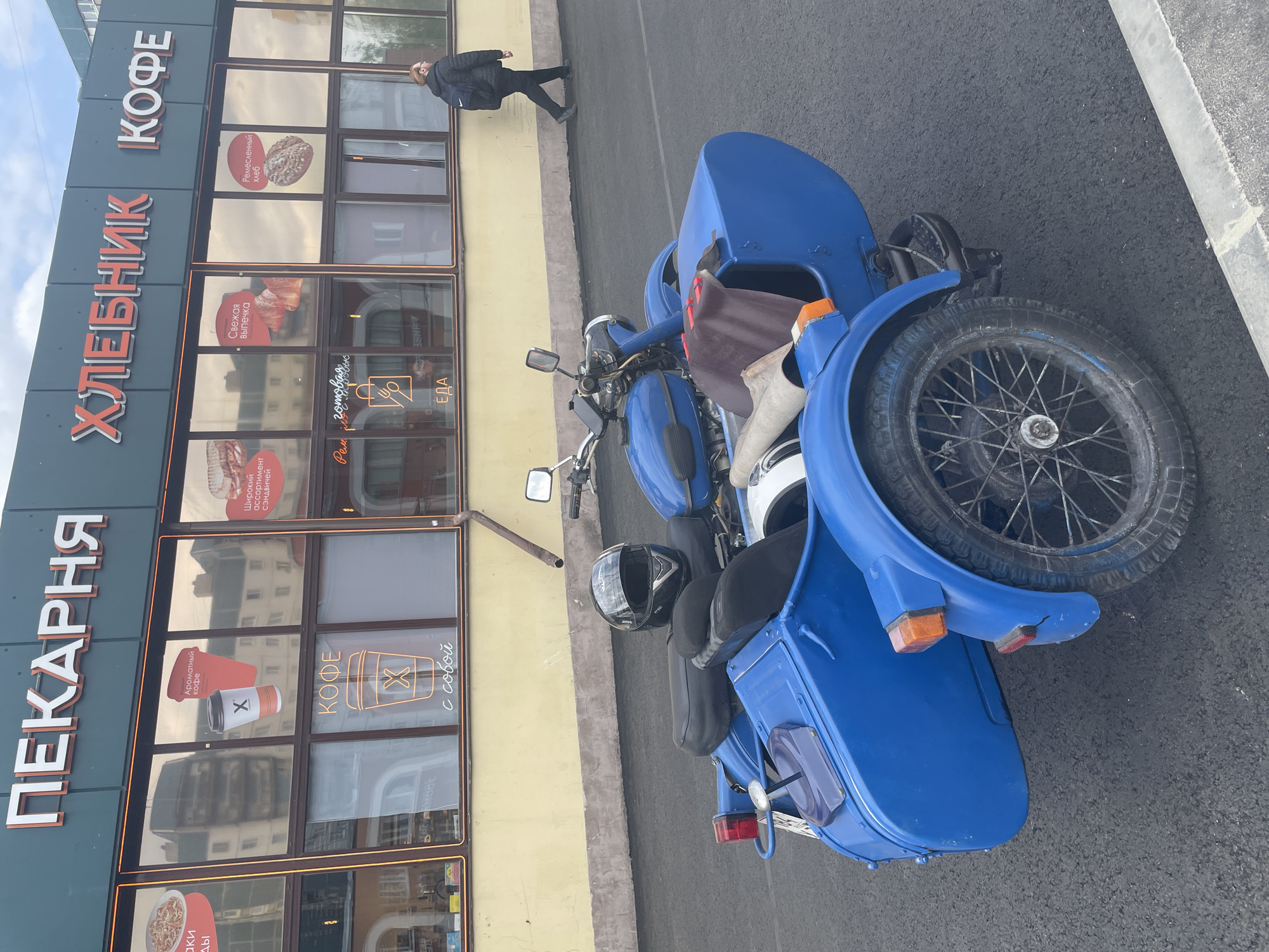 Here's a detailed description of the image:
**Scene:** The image shows a bright blue motorcycle with a sidecar parked on an asphalt street in front of a building.
**Location:** The building appears to be located in Saint Petersburg, Russia, based on the Cyrillic script on the storefront. The sign reads "Пекарня Хлебник Кофе", which translates to "Bakery Khlebnik Coffee" and displays images of baked goods and coffee.
**Objects and Subjects:**
* A blue motorcycle with a sidecar is prominently featured. Two helmets sit on the motorcycle.
* The sidecar has a cover and a spare tire.
* A pedestrian dressed in dark clothing is walking along the sidewalk by the building.
**Environment:**
* The street is asphalt, showing typical wear and tear.
* The building has large windows displaying advertisements for the bakery/coffee shop. The facade has a combination of gray, yellow and black paneling.
**Additional details discernible from the storefront signs (translated):**
* "Fresh Pastries"
* "Handcrafted Bread"
* "Wide Assortment of Sandwiches"
* "Aromatic Coffee"
* "Coffee to Go"
* "Ready-made food"