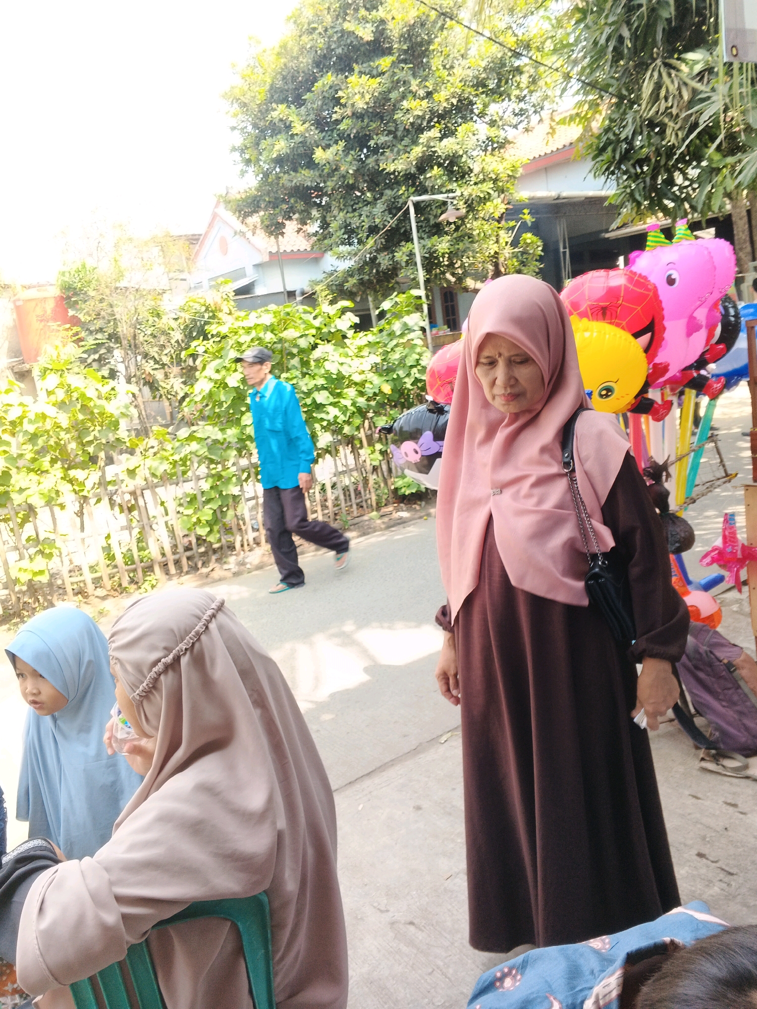 In Kadu, Indonesia, an outdoor scene unfolds under bright sunlight. In the foreground, a woman wearing a brown abaya and a dusty pink hijab stands looking downwards, a black purse with a chain strap slung over her shoulder. To her left, a young child in a light blue hijab is partially visible, seemingly engaged in conversation with another person out of frame. Behind them, a man in a bright blue long-sleeved shirt and dark pants, wearing a black cap, walks away from the viewer. He is on a paved surface, with a bamboo fence and lush green foliage to his left. In the background, colorful balloons, including a pink pig and a yellow round balloon with a face, are displayed on a stand, suggesting a local market or event. Buildings with red-tiled roofs are visible behind the trees. The overall impression is a snapshot of daily life in a tropical urban environment.