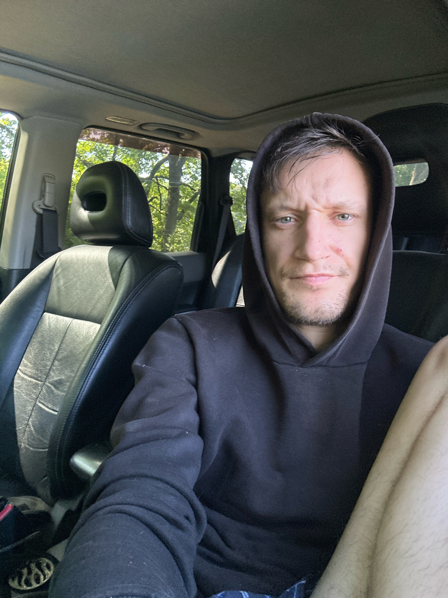A close-up, over-the-shoulder selfie shows a man with light brown hair, light blue eyes, and a small scar on his right cheekbone, wearing a black hooded sweatshirt. He has a faint beard and mustache. He is seated in the driver's seat of a car, with the passenger seat and headrest visible to his left. The interior of the car is dark, with a grey headliner and a sunroof. Sunlight streams in through the windows, illuminating trees and foliage outside. The man's left leg is visible in the lower right portion of the image. The scene appears to be set in a wooded area, possibly on a road or in a park, in or near Makiv, Ukraine.