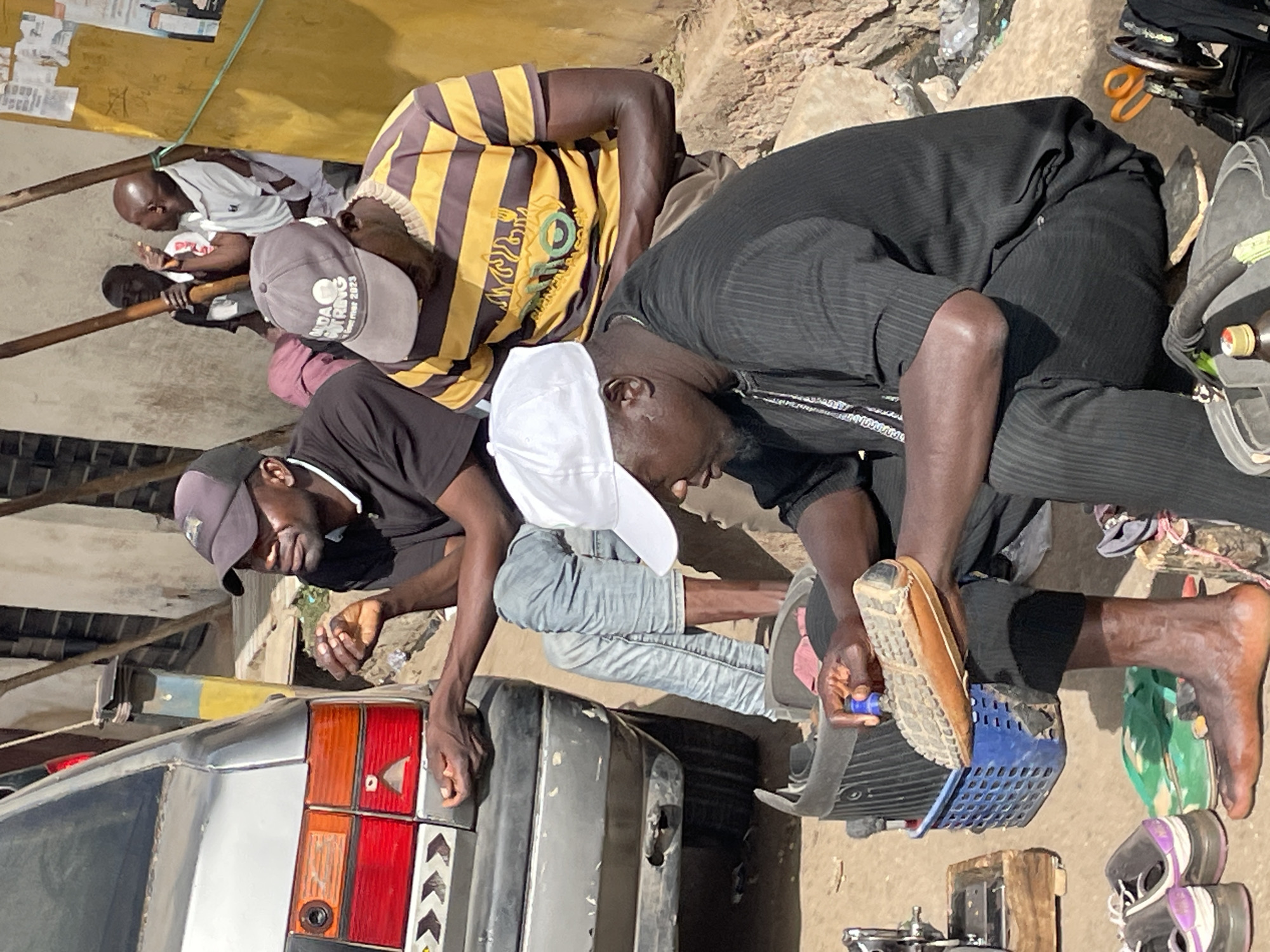 The image depicts an outdoor scene in Jos, Nigeria, where several men are gathered around the rear of a silver car, suggesting an informal repair or inspection activity. At least six men are visible, most of whom are bending over the vehicle's taillights and bumper.
In the immediate foreground, a man in a black t-shirt and dark baseball cap leans over the left rear of the car. Adjacent to him, another man, wearing a white baseball cap and light blue jeans, is also crouched down. To the right, a man dressed in a black striped shirt and black trousers is deeply crouched; one foot is bare while the other, clad in a brown shoe, rests on a blue plastic basket. He is holding a small blue object, possibly a tool. On the dusty ground near him are a pair of green flip-flops, purple sneakers, orange scissors, and a small bottle with a yellow cap.
Further back, a man in a yellow and brown striped t-shirt and a gray baseball cap is also leaning towards the car. The gray cap has discernible text: "UDA PUT RING" and "2023". Behind him, partially visible, is a bald man wearing a white t-shirt. The background features rough concrete blocks, dirt ground, and simple structures that appear to have posters or papers attached, indicating a casual, possibly roadside setting. The bright, clear lighting suggests it is daytime. The men appear engaged and focused on the task at hand. The car's rear bumper seems to be dislodged or undergoing work.