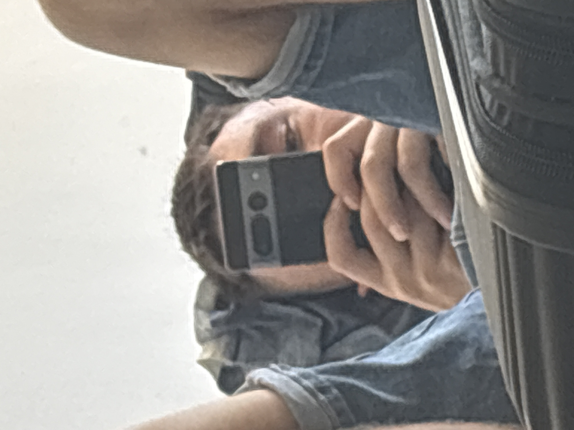 A person is lying on their back and holding a smartphone up to their face, obscuring most of their features. They are wearing a denim jacket and what appears to be jeans. The background is a plain white wall. The person's legs are raised and spread apart, and their arms are bent as they hold the phone. The scene appears to be indoors, possibly in a home or hotel room.