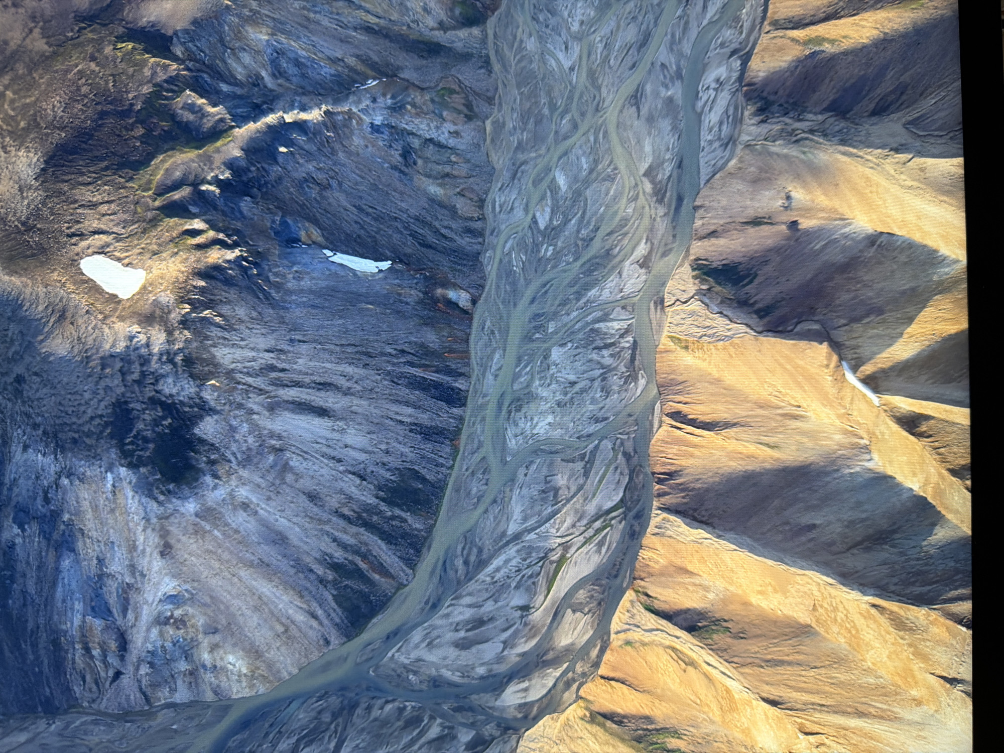 This is an aerial photograph of a mountainous, glacial landscape. The terrain features steep, rocky slopes in shades of blue, gray, and brown, with patches of green vegetation and areas of snow or ice. A braided river system, appearing as multiple winding channels of light blue-green water, flows through a valley. The riverbeds are composed of light-colored sediment. The foreground is dominated by a sunlit, golden-brown mountain range with dramatic shadows cast by the rugged topography. The image does not depict any urban environment or specific location within Kyiv, Ukraine, as it portrays a natural, remote landscape.