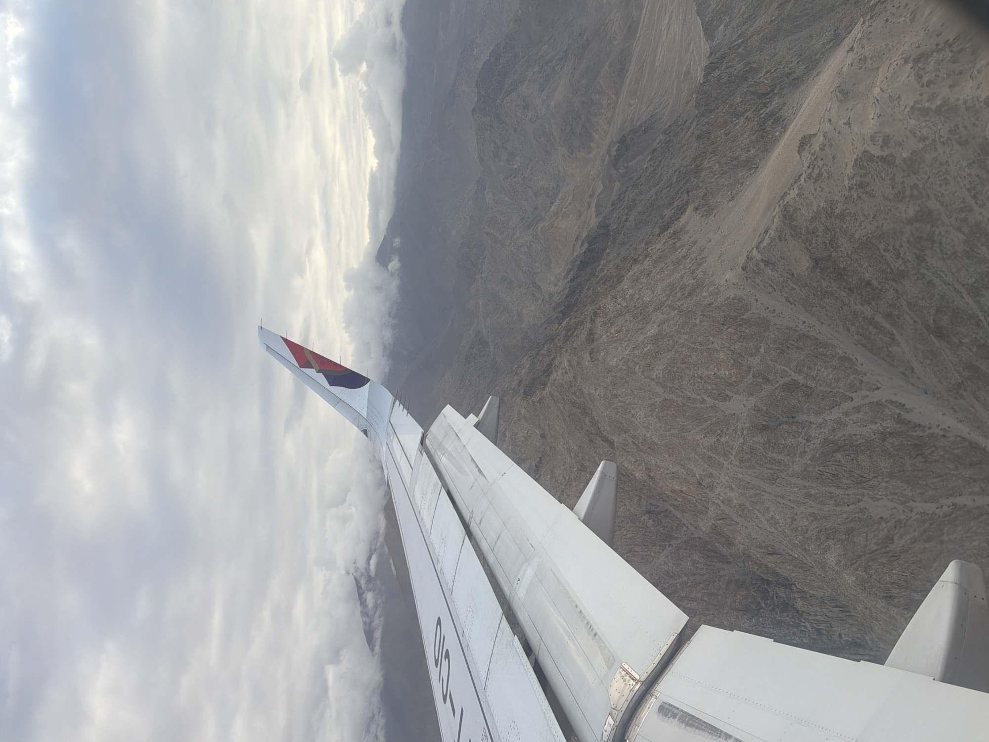 An aerial view from an airplane reveals a mountainous landscape under a cloudy sky. The right side of the image is dominated by rugged, barren mountains with visible geological formations. A portion of the airplane's wing, white with some dark lettering, is prominent in the foreground, extending from the left edge of the frame towards the center. The wing has visible flaps and mechanical components. The tail fin of the airplane, featuring a colorful logo, is visible at the top right corner. The clouds are scattered, with some obscuring the peaks of the distant mountains. The scene appears to be over a high-altitude, arid region, consistent with the geography around Leh, India. There are no people or vehicles visible in the image.
